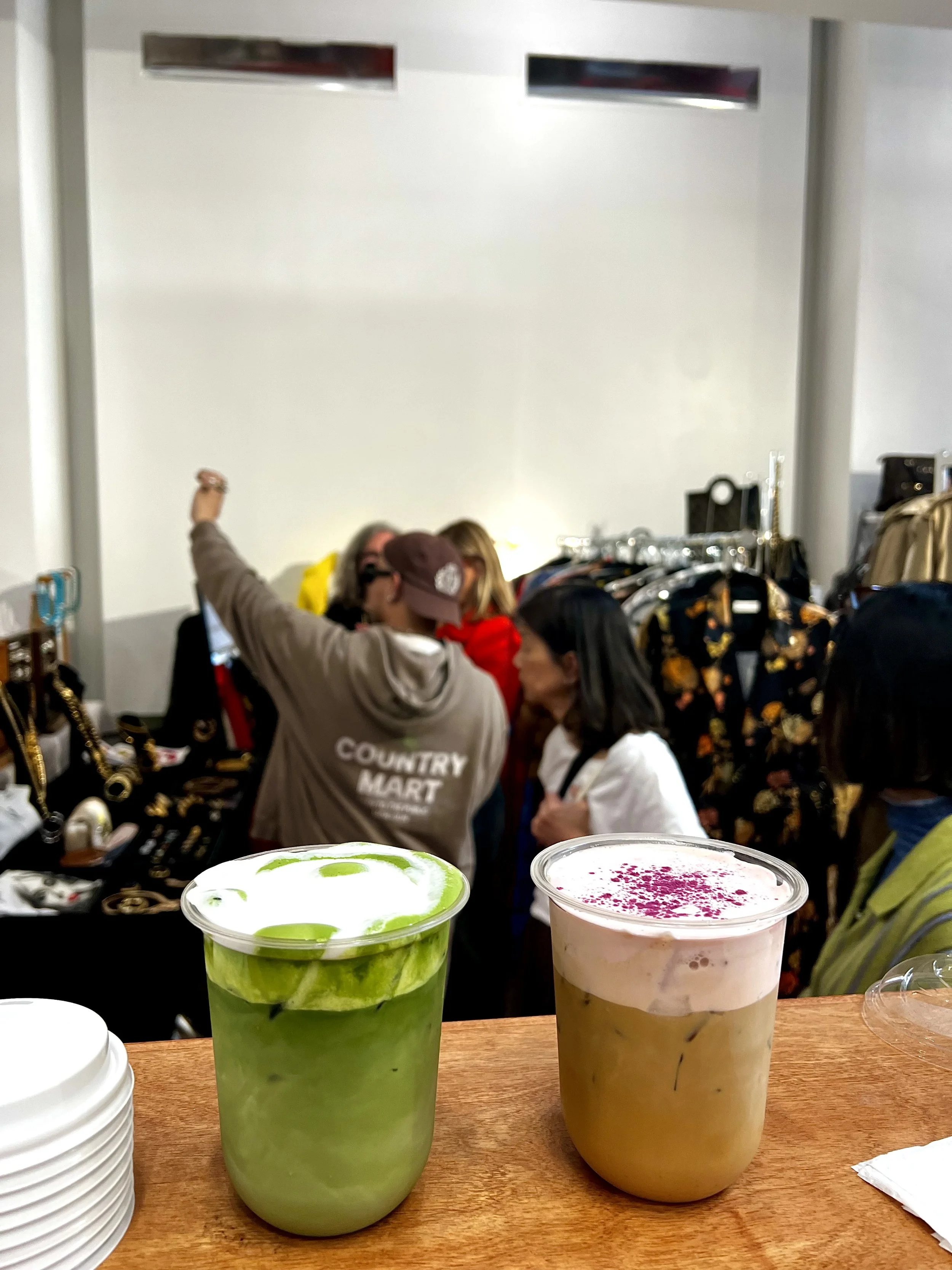 Two beverages on a wooden table, one green and one pink with purple sprinkles, in a busy marketplace with people and clothing racks in the background.