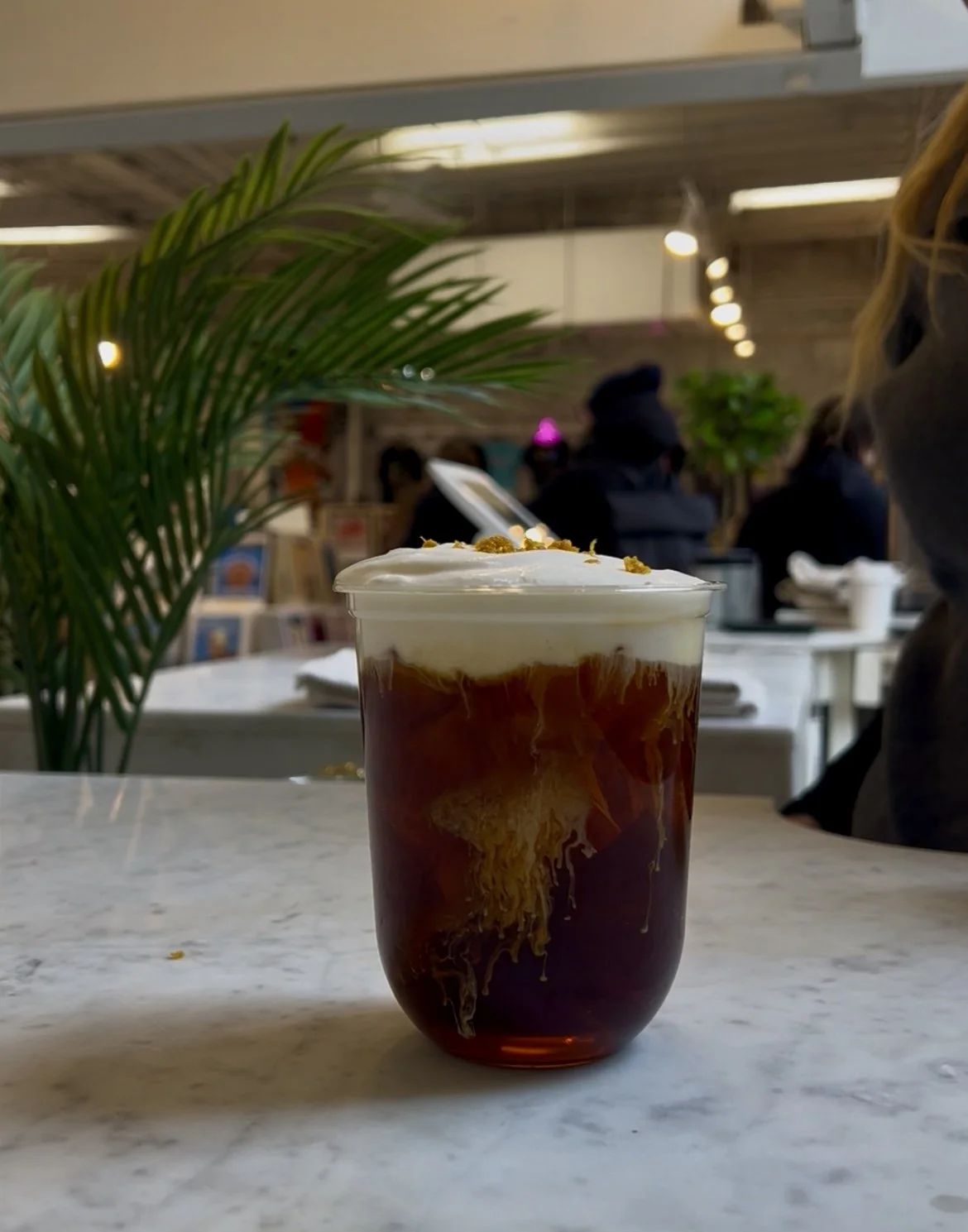 A cold coffee beverage in a glass with swirls of cream or milk, topped with whipped cream and small garnish, placed on a marble table in a cafe setting. Mont Blanc, strong cold brew with orange cold foam