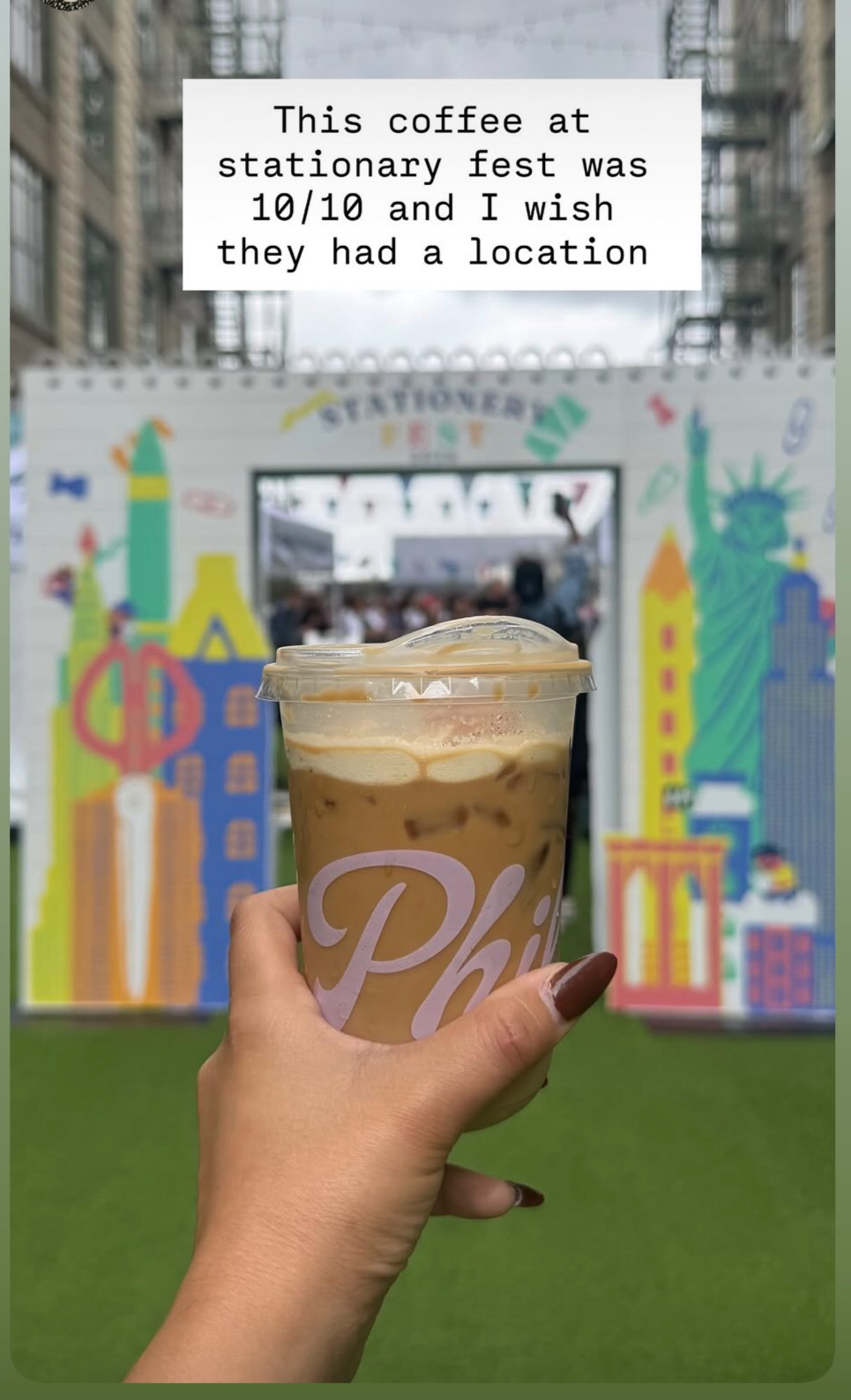 Hand holding iced coffee in front of a colorful backdrop with a sign that says "Stationery Fest" at a festival or outdoor event.