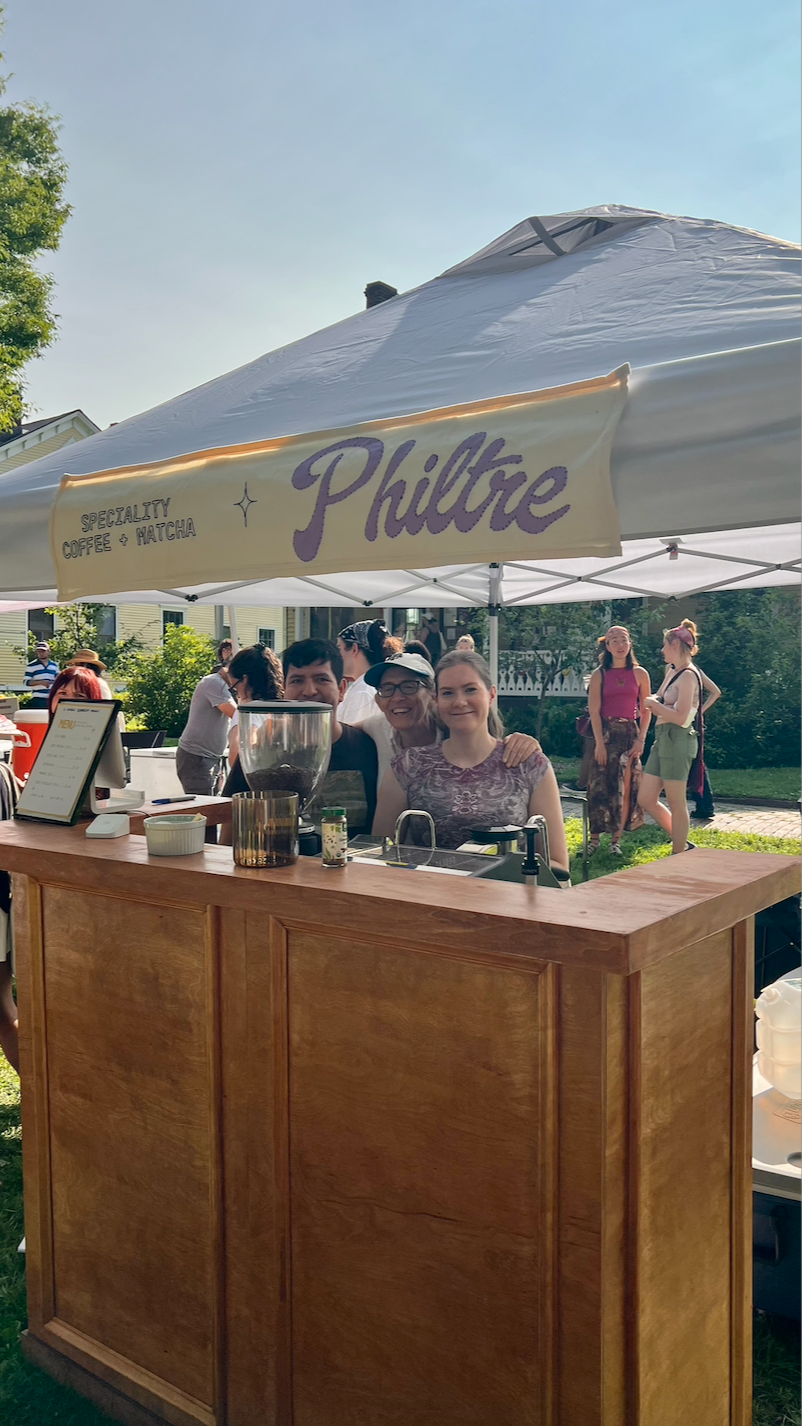 People at a coffee stand under a canopy with a sign that reads "Philtre" and mentions specialty coffee and matcha, outdoor setting, sunny weather, trees and houses in the background.