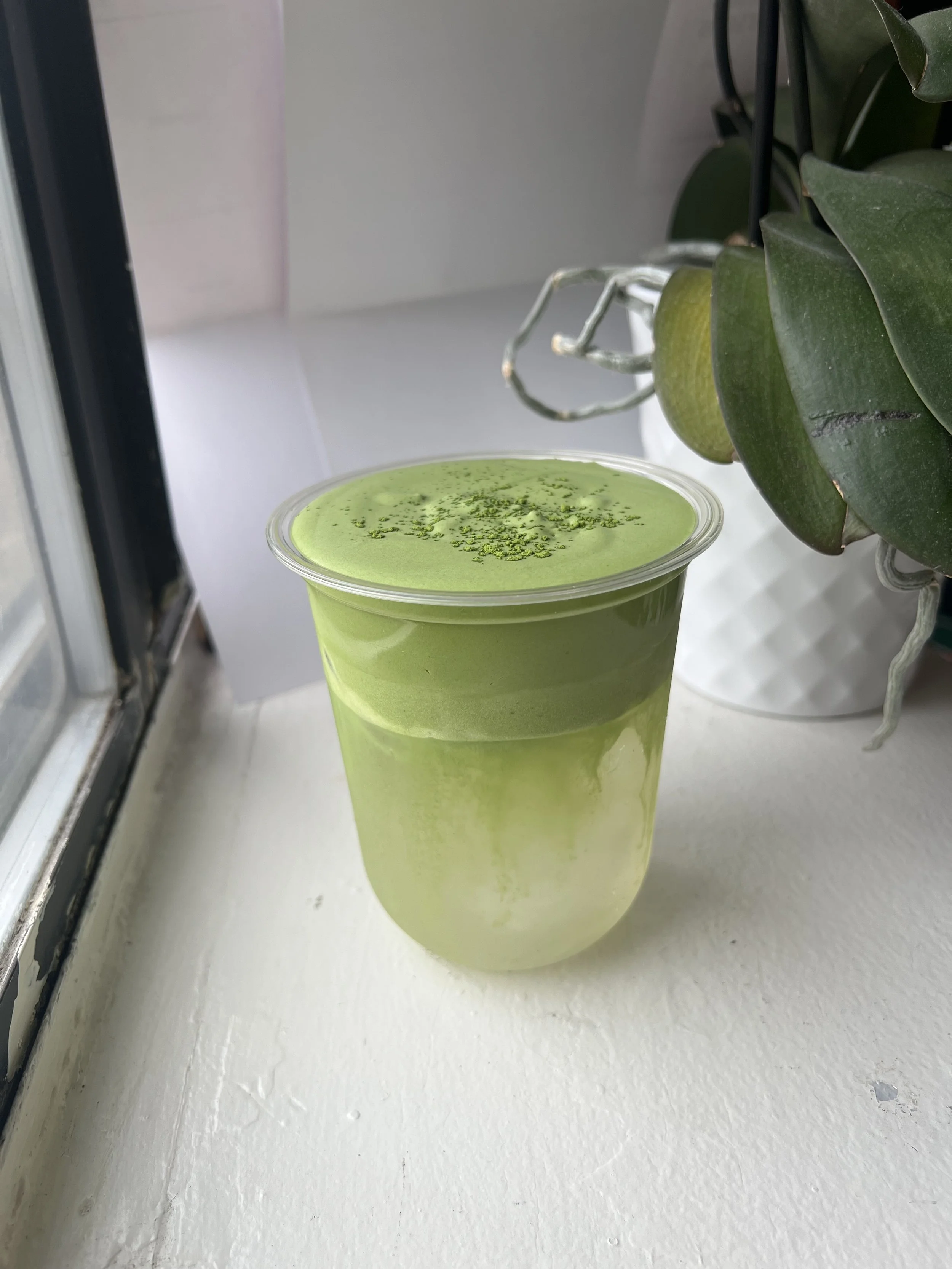 A clear plastic cup filled with a layered green beverage, placed on a white windowsill near a potted plant with dark green leaves. Coconut matcha cloud.