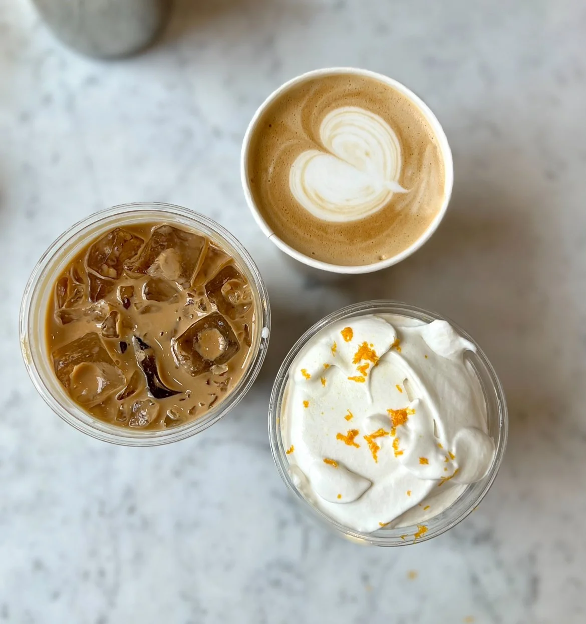 Overhead view of three beverages on a white surface: iced coffee with ice cubes, a latte with heart-shaped foam art, and a drink topped with whipped cream and orange zest. Speciality, elegant coffee experience.