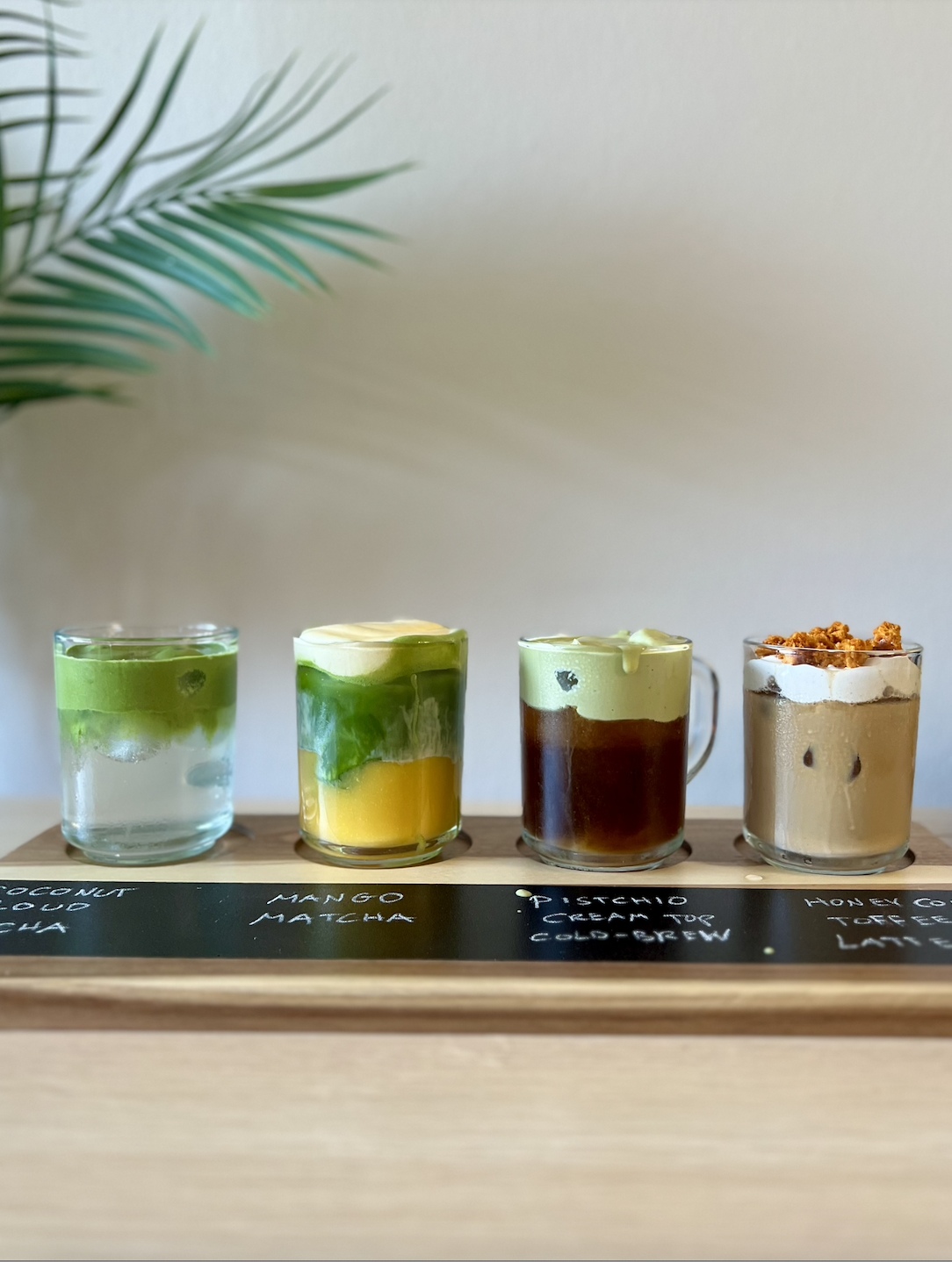 Four transparent coffee cups with different layered drinks placed on a wooden tray, with a blackboard label underneath each drink. A green plant in the background. 4 speciality drink flight