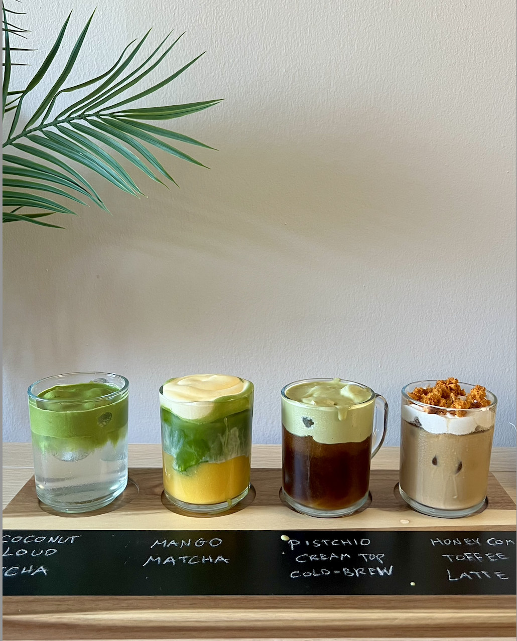 Four transparent glasses each filled with different layered drinks, placed on a wooden tray with handwritten labels underneath. The first drink has green jelly or fruit pieces in a clear liquid, the second has a yellow base topped with green jelly, the third contains a dark liquid with green cream on top, and the fourth has a layered coffee drink topped with whipped cream and crunchy topping. A plant with long green leaves is partially visible in the top left corner.