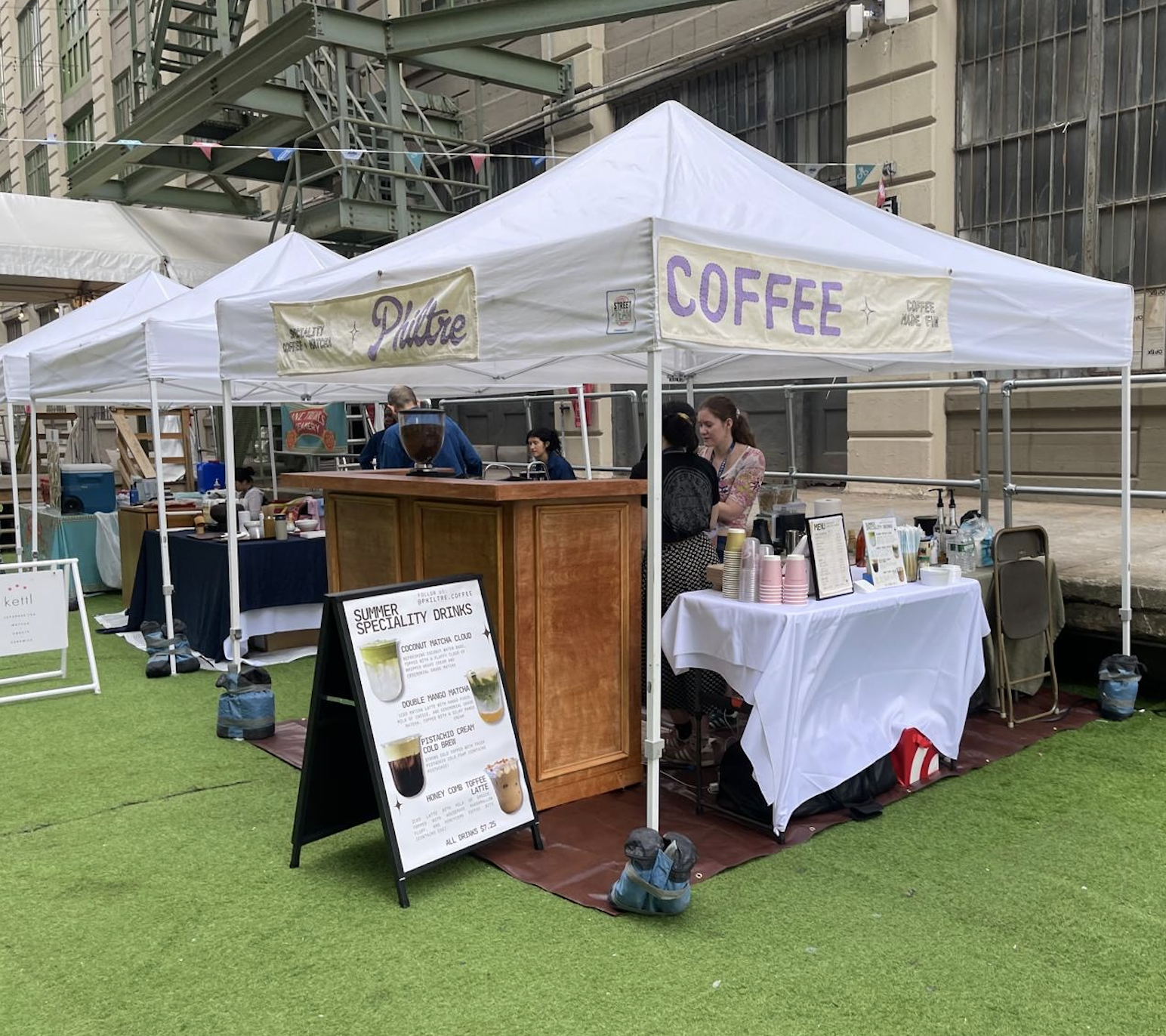 A coffee set up at industry city with a white canopy offering coffee and summer specialty drinks, with signs and tables displaying cups, menus, and equipment, set on a grassy outdoor area surrounded by urban buildings.
