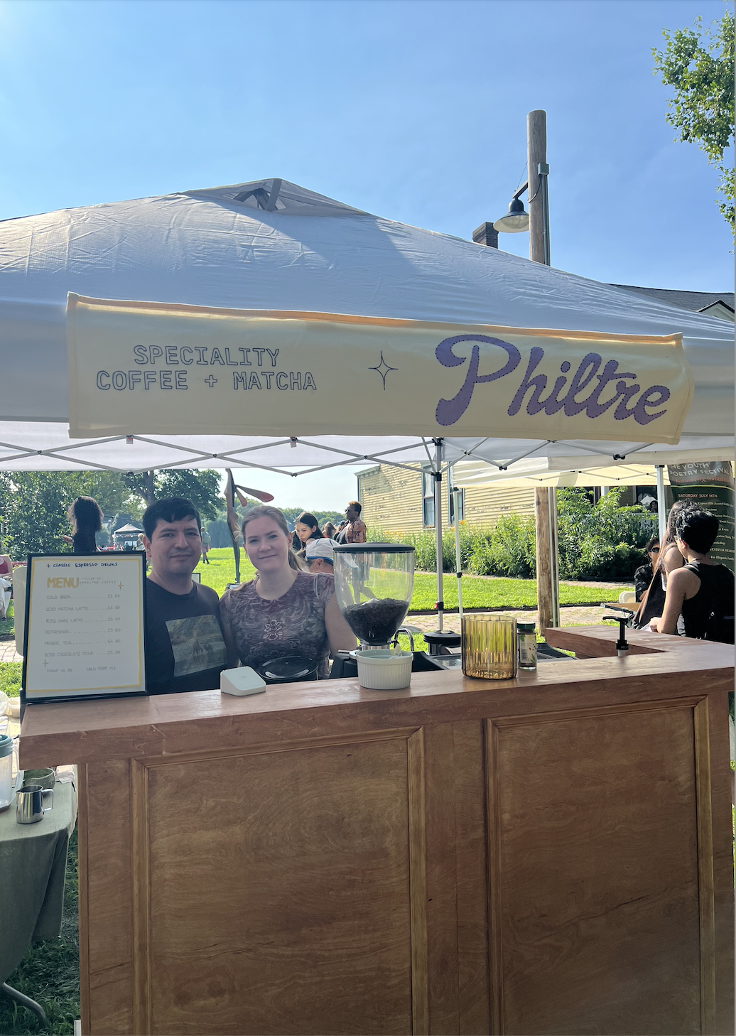 A coffee stand under a white tent with a sign that reads 'Specialty Coffee + Matcha Philtre.' Two people are standing behind the counter, with a menu and coffee equipment visible, outdoors at a sunny event.