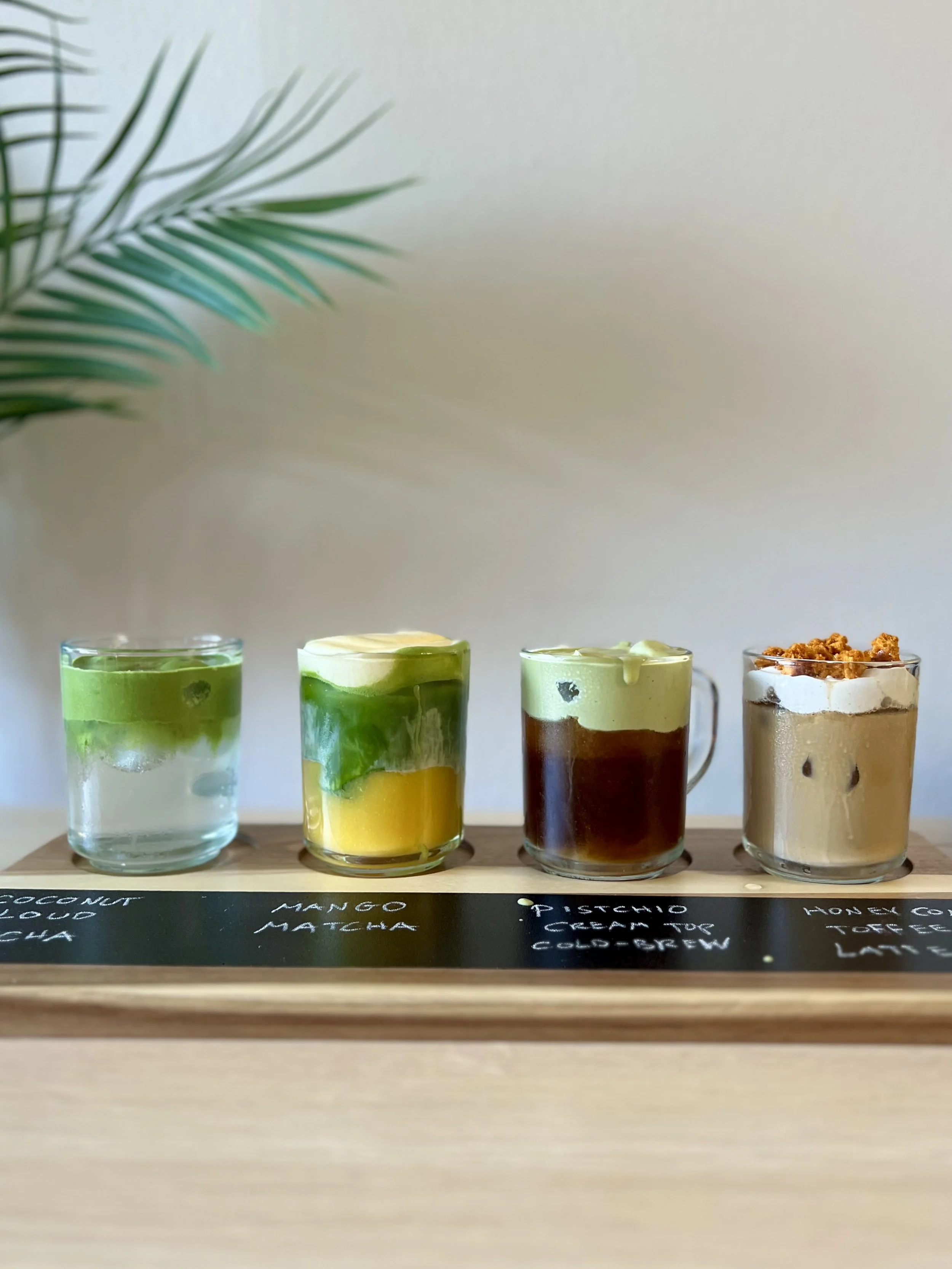 Four colorful layered drinks in clear glasses on a wooden tray, with a chalkboard menu below and a green plant in the background.