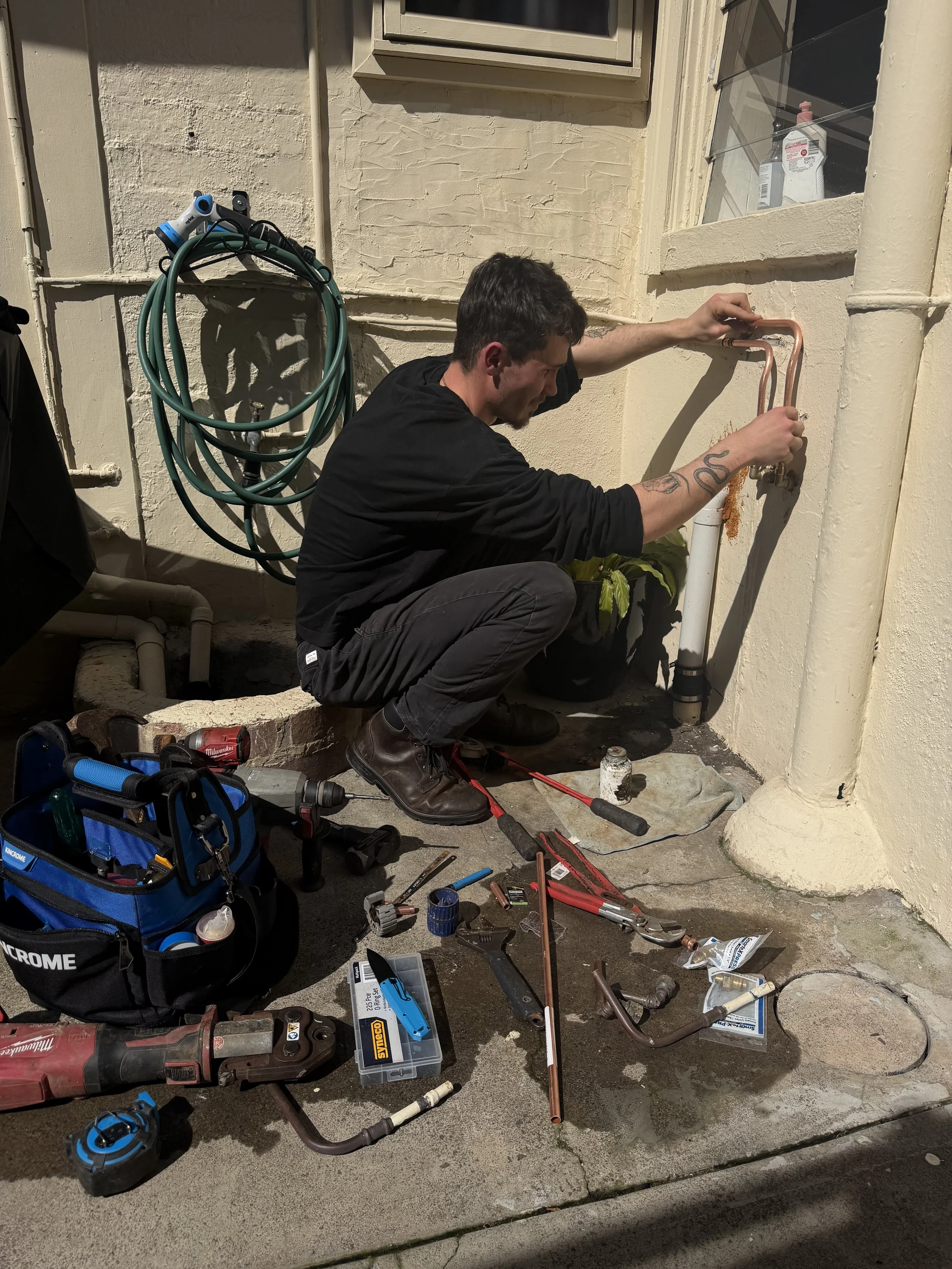Man working on outdoor plumbing pipes, surrounded by tools and equipment, in front of a beige wall with a window and a plant.