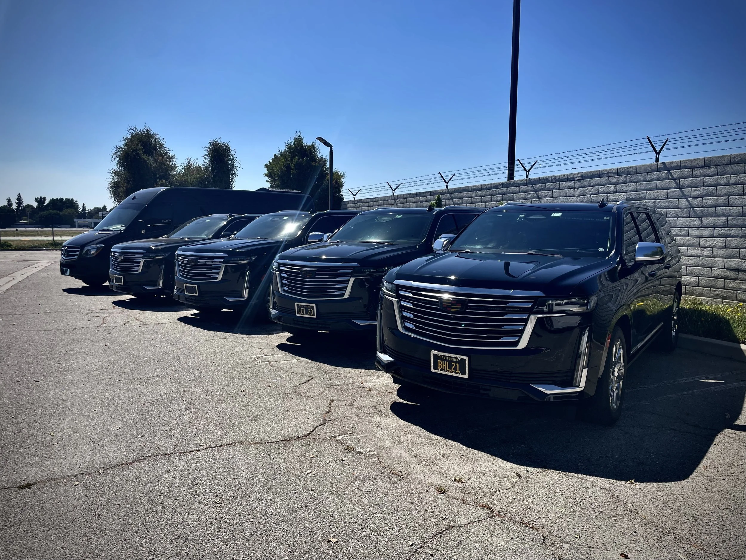 Several black luxury cars, including a Cadillac, parked in a lot near a stone wall topped with barbed wire, under a clear blue sky.