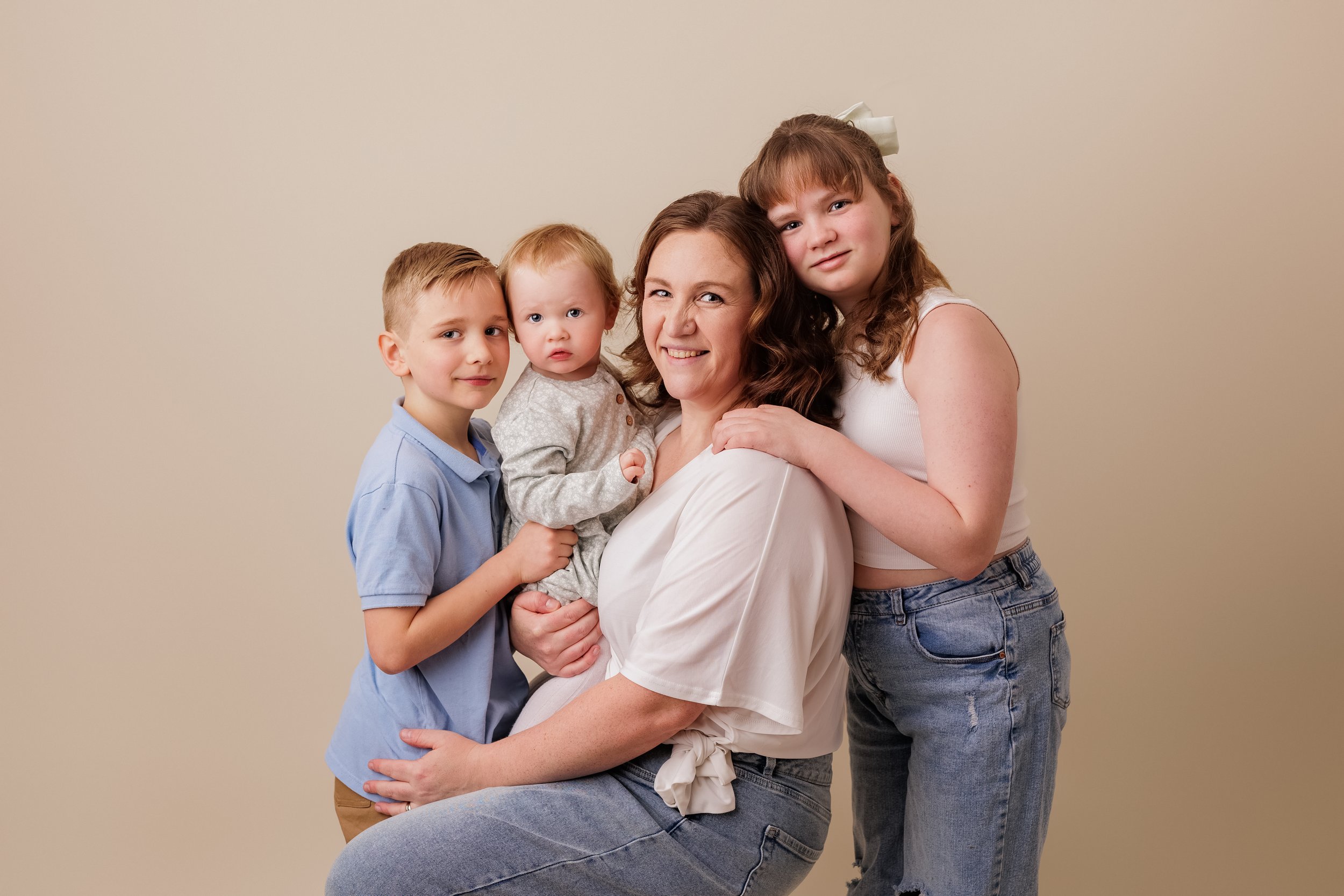 Cute family photo with a smiling mother and three children, all dressed casually, posing against a plain beige background.