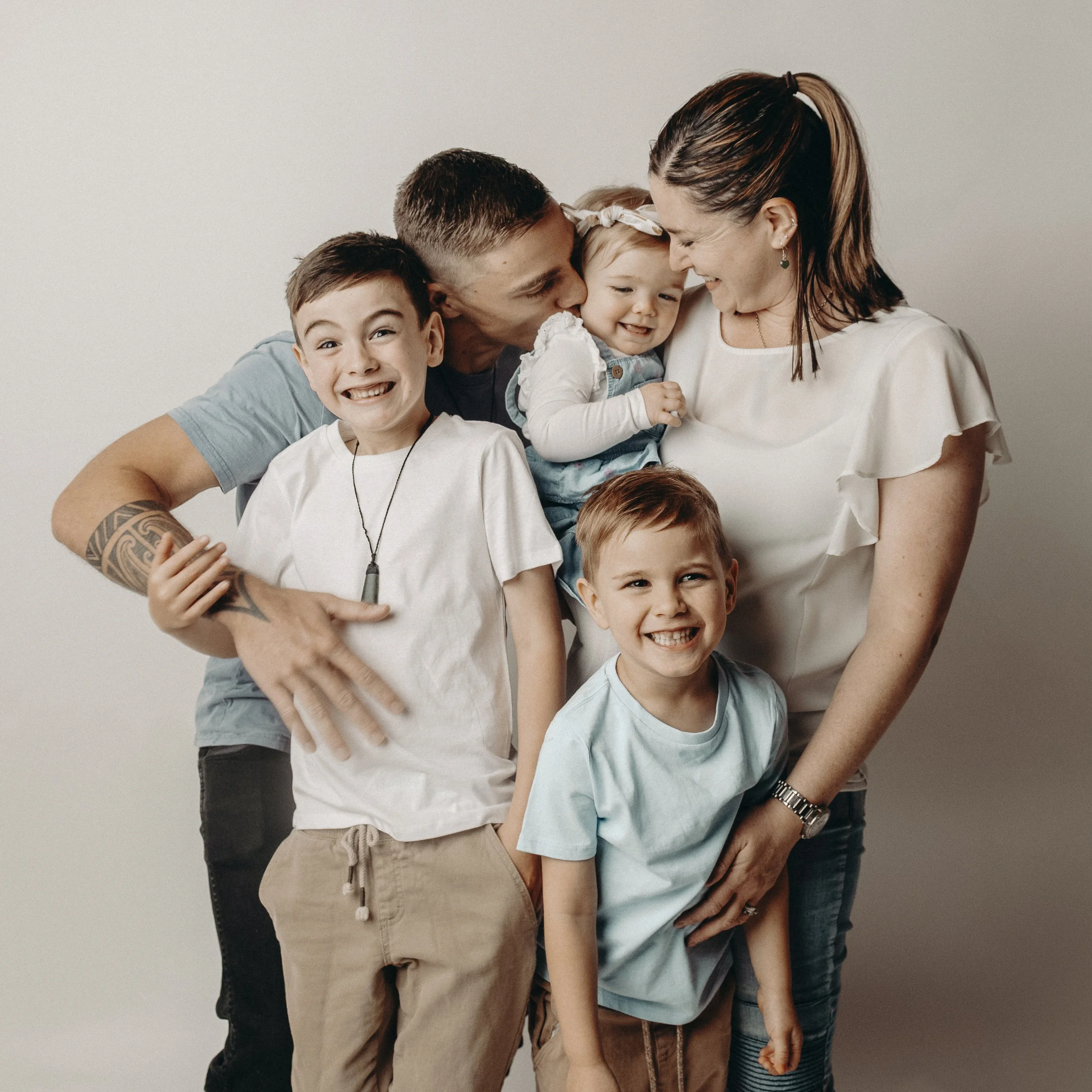 A family of six, including two children and two adults, sharing an affectionate moment, with one of the adults kissing the youngest child on the cheek, against a plain background.