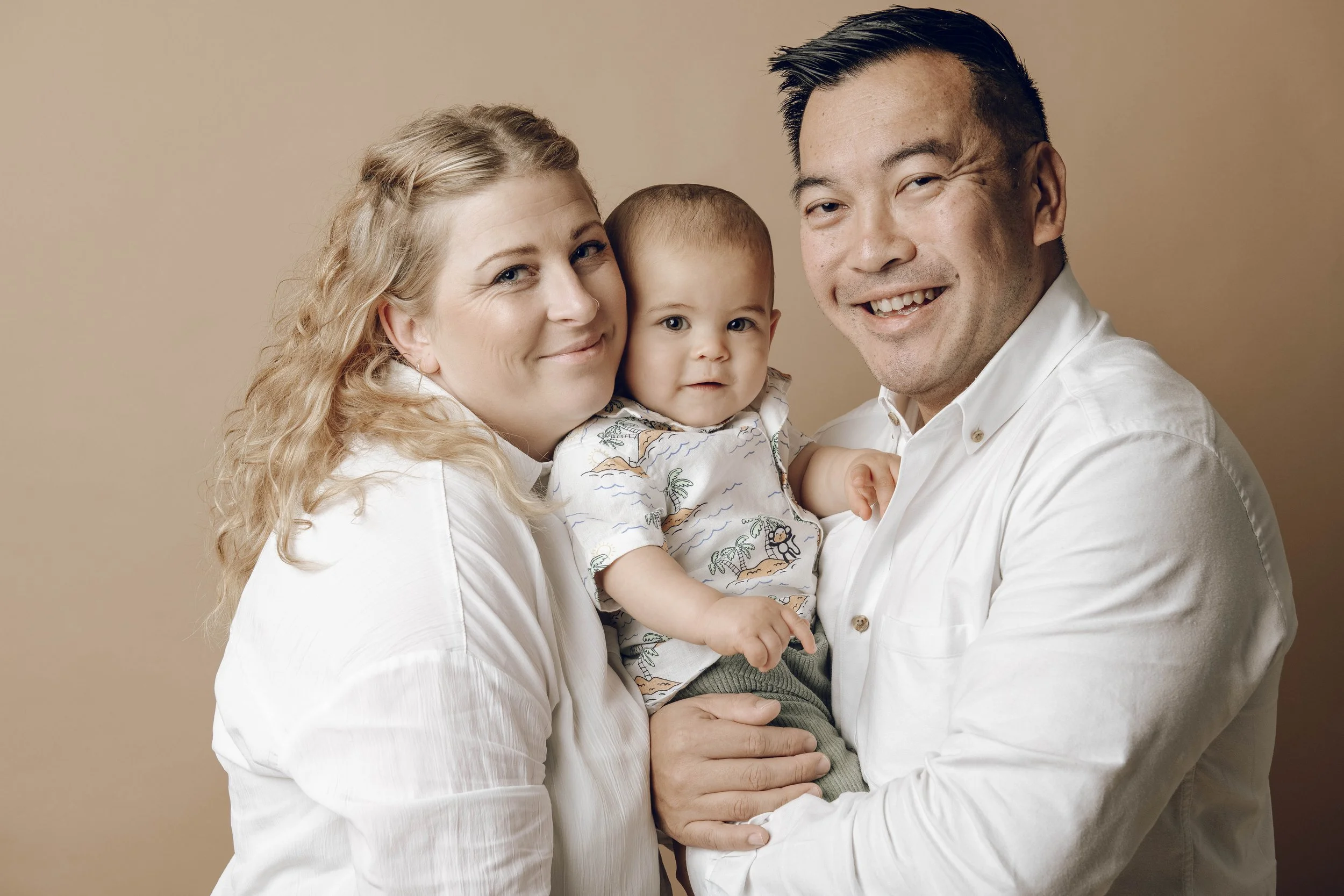 A smiling family of three, including a woman with long, curled blonde hair, a man with short black hair, and a young boy with short hair, all wearing white tops, against a beige background.