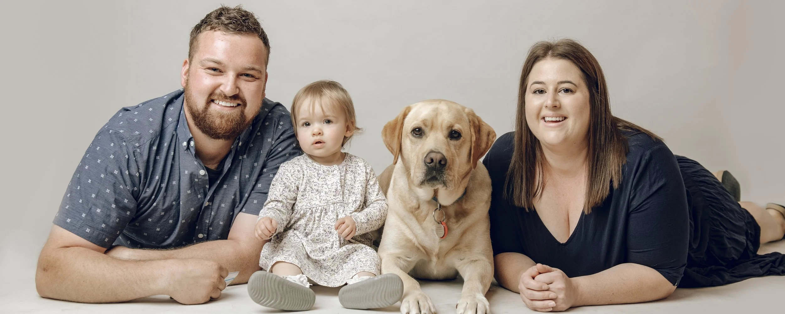 A family of four with a dog lying on the floor, all smiling and looking at the camera against a plain background.