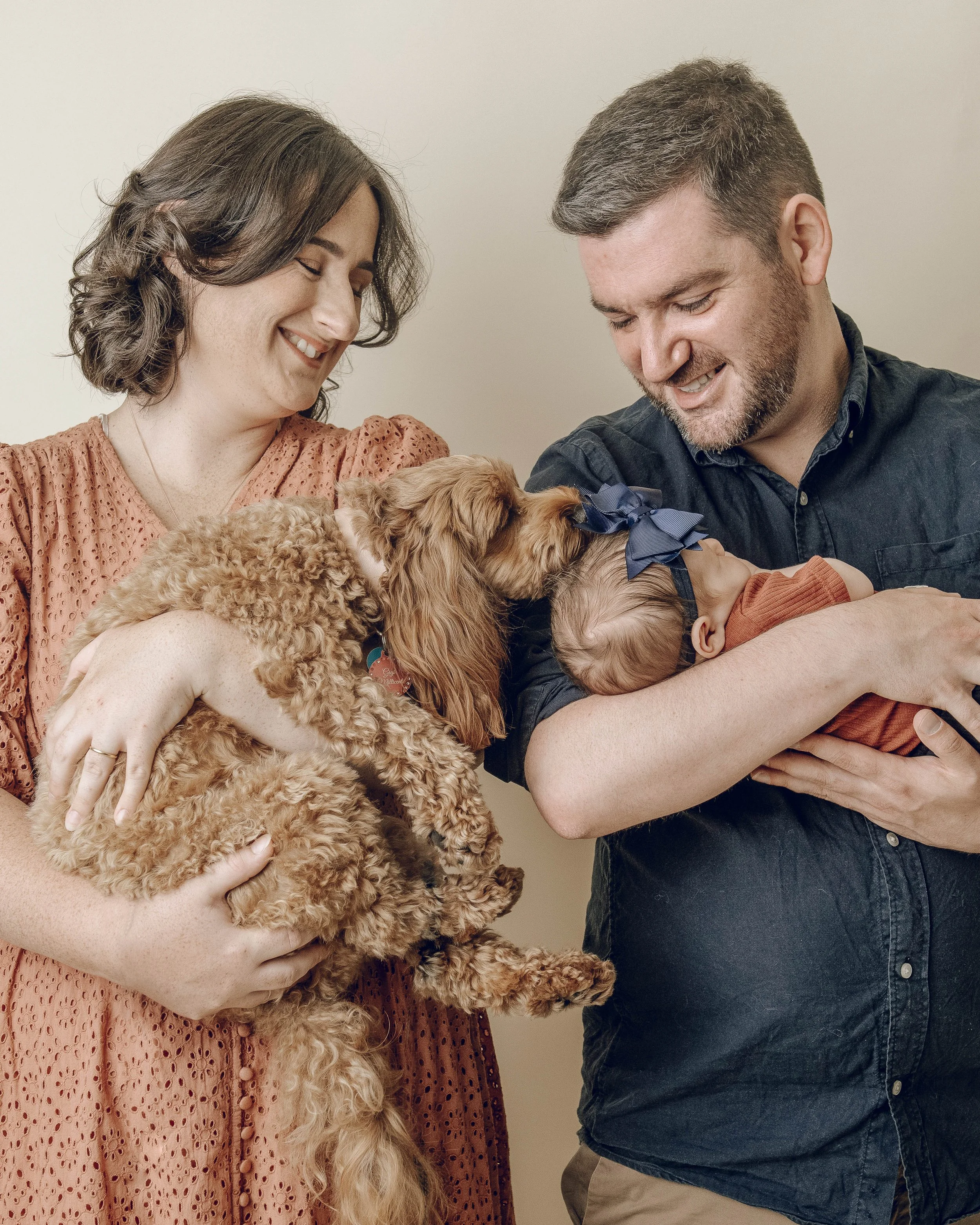 A man and woman smiling and holding a fluffy brown dog with a blue bow, in front of a plain beige background.