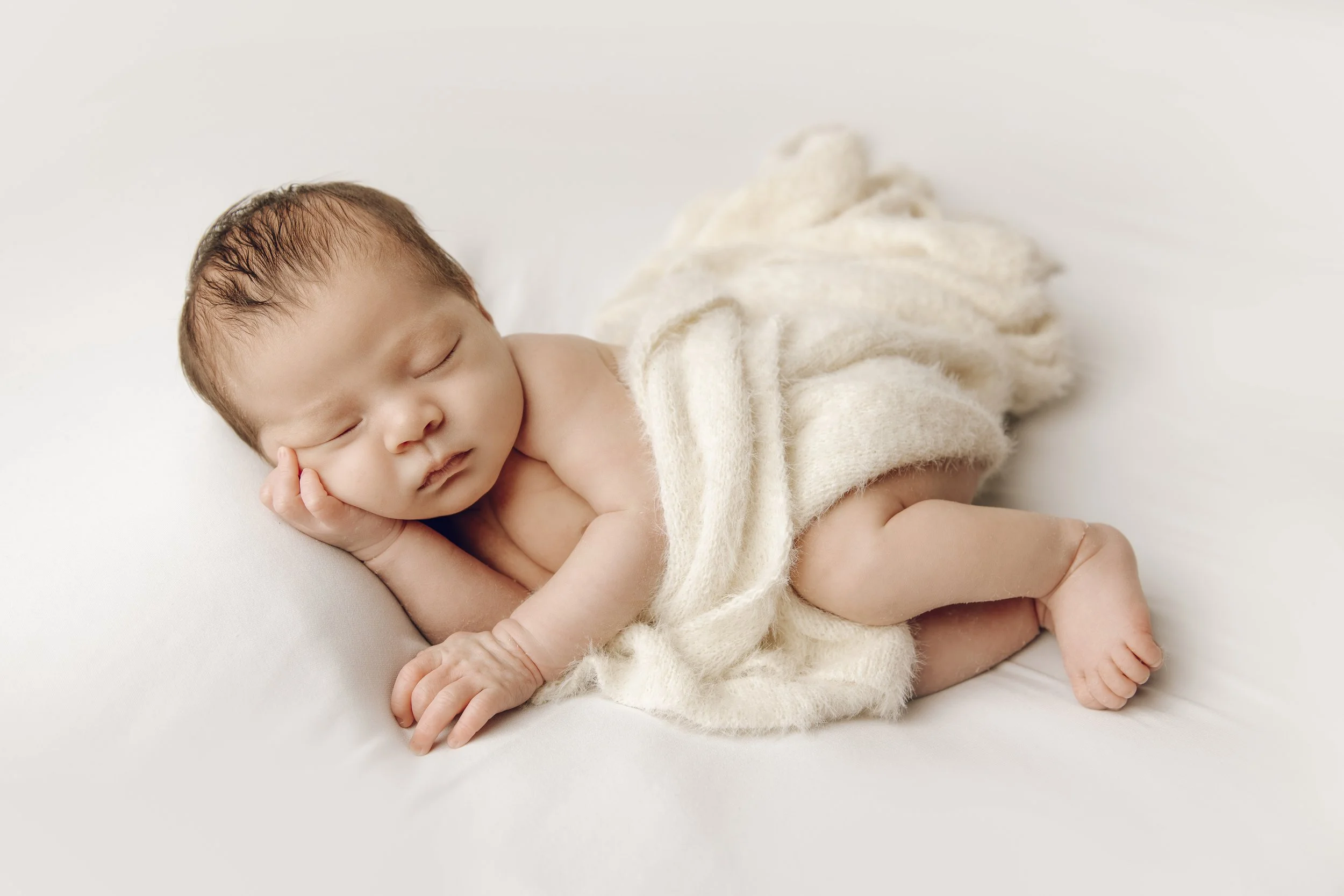 A sleeping baby with brown hair, lying on its side on a white surface, wrapped in a soft cream-colored blanket.