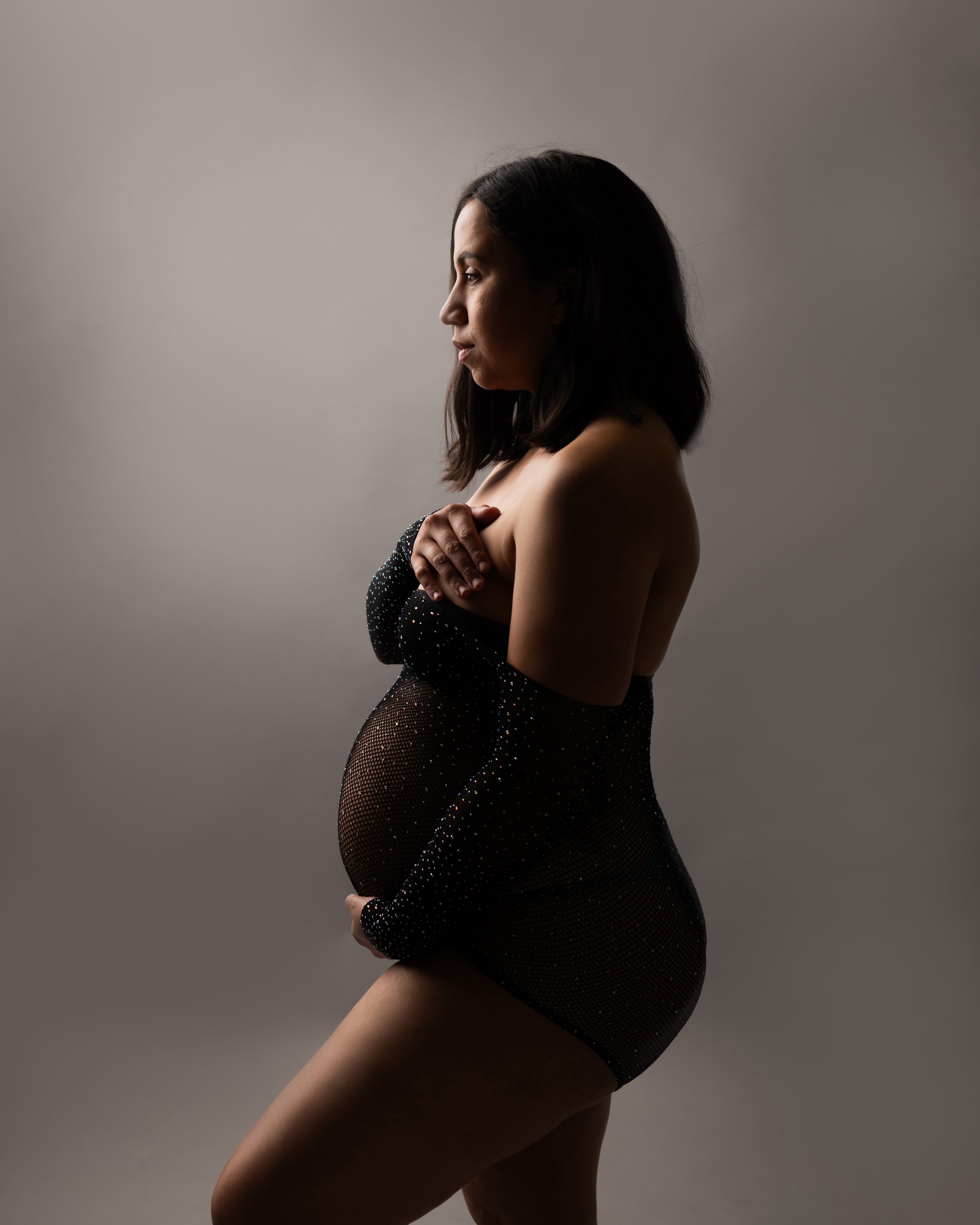 Profile view of a pregnant woman with shoulder-length dark hair, wearing a black dress with sparkles, standing against a plain gray background.