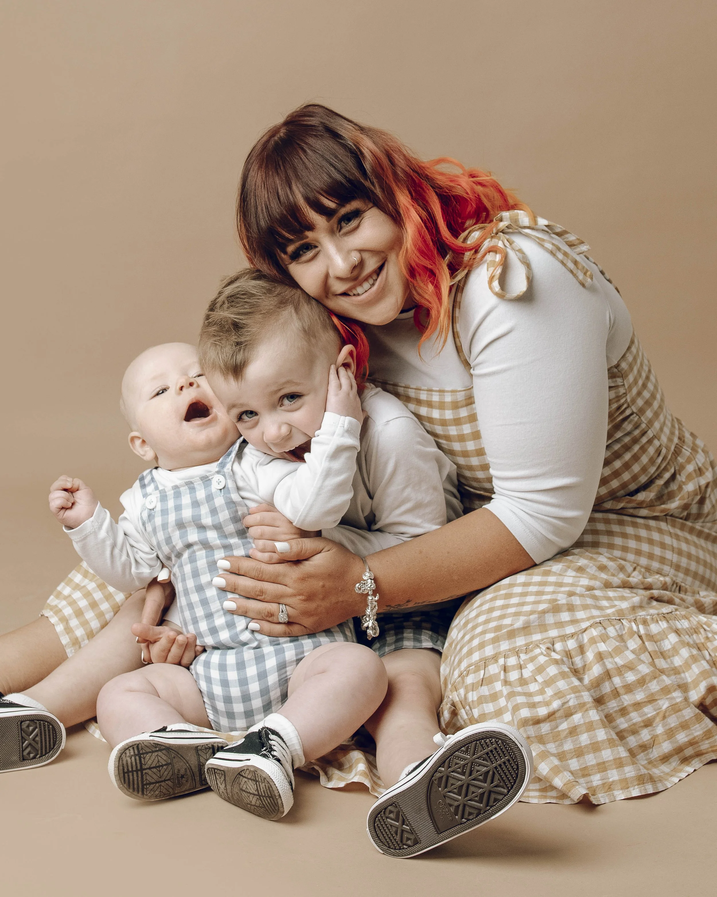 A woman with bright orange and red hair, smiling and hugging two young boys, one of whom is bald and the other has light brown hair. They are all sitting on the floor against a beige background. The woman is wearing a beige and white checkered dress 