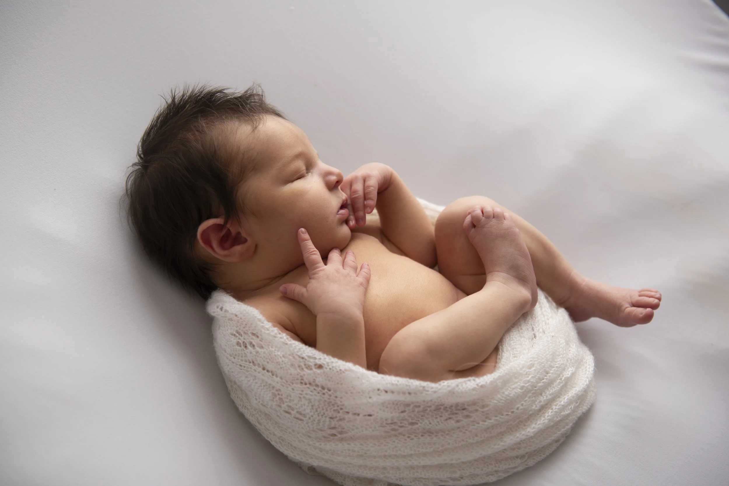 A sleeping baby with brown hair wrapped in a soft white blanket, lying on a white surface.