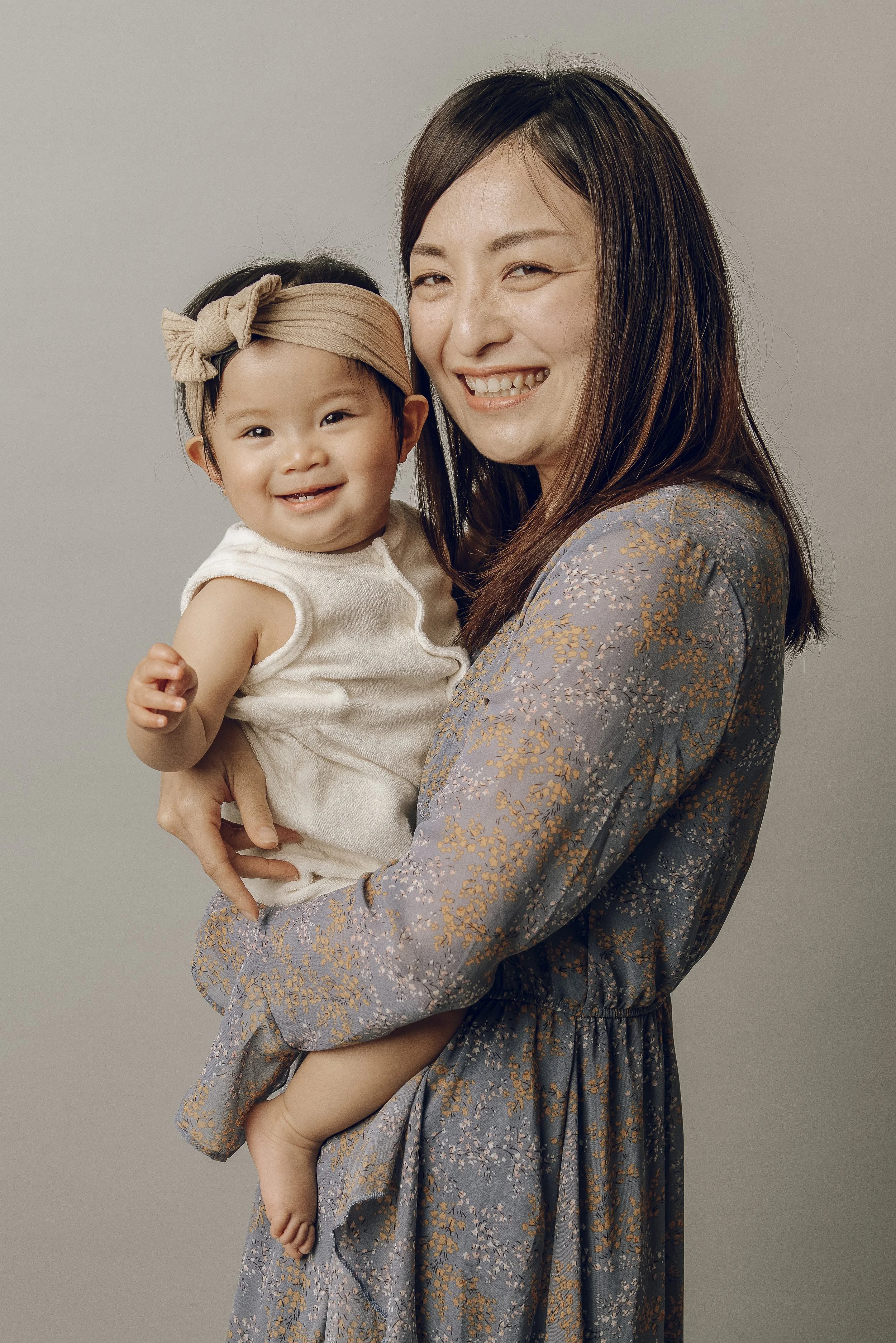 A woman holding a smiling toddler girl against a plain gray background.