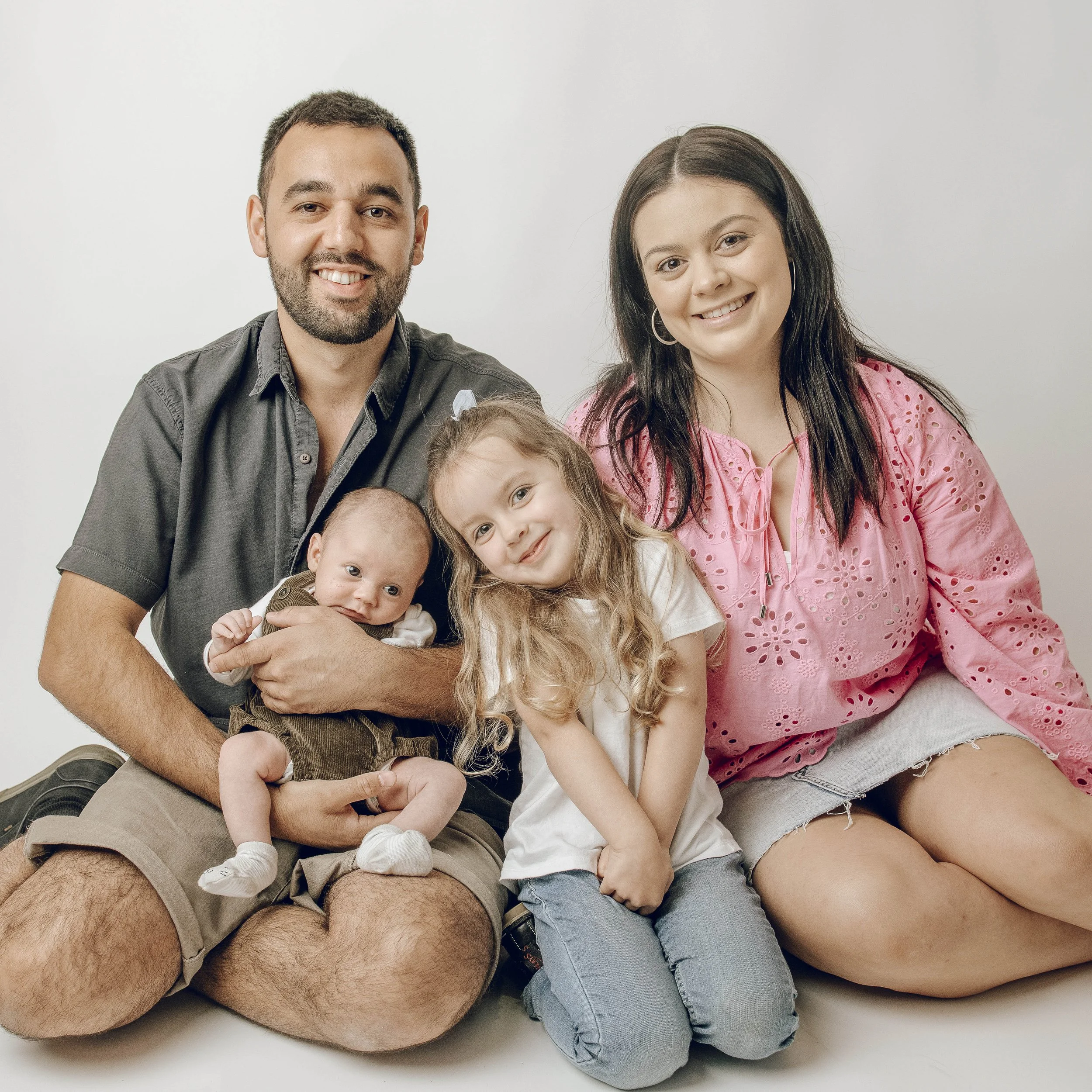 Smiling man and woman sitting close together with three young children, all smiling, against a plain white background.