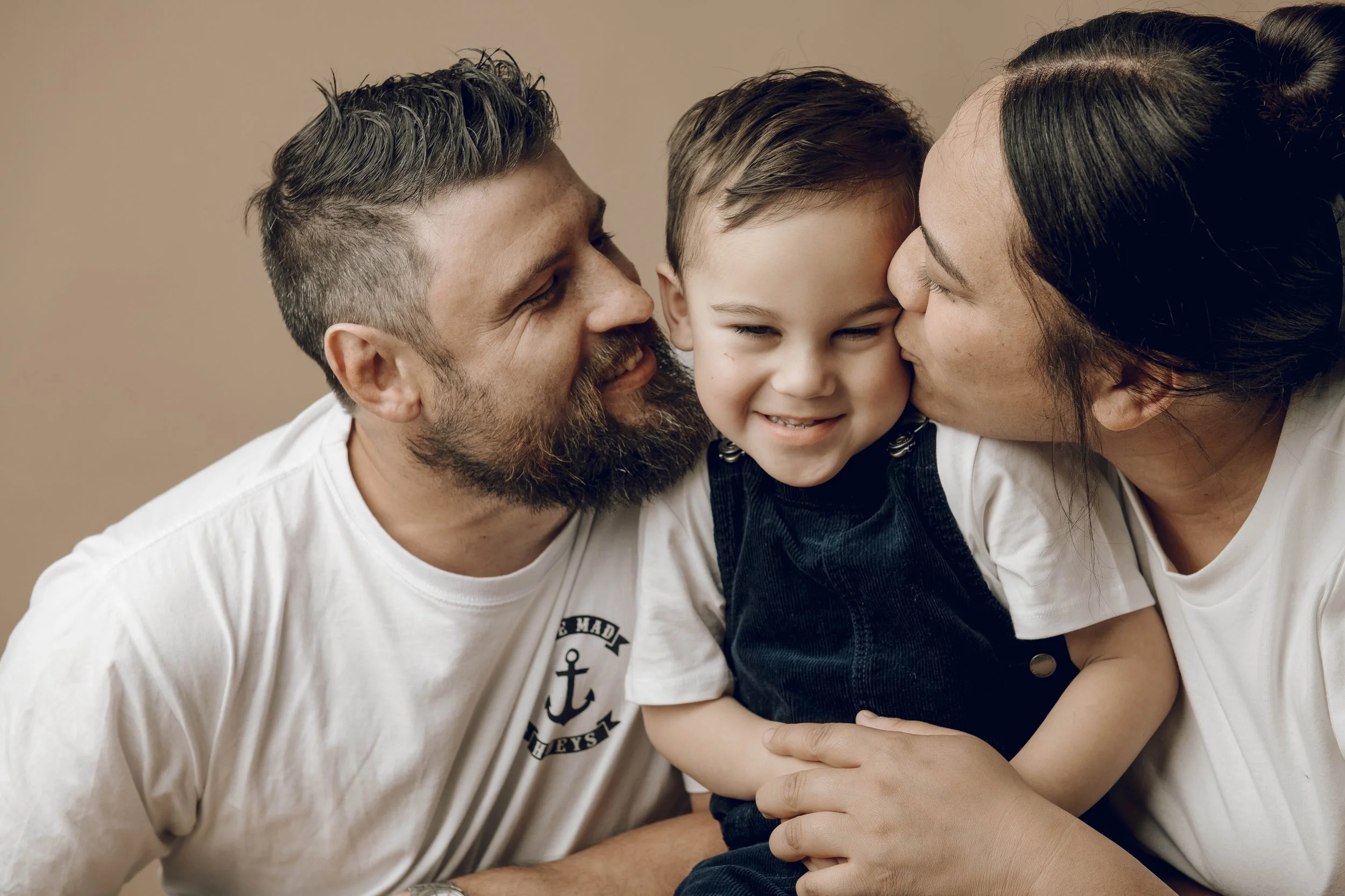 A family of three, a man, a woman, and a young boy, share a loving moment close together, with the woman and man kissing the boy's cheeks.
