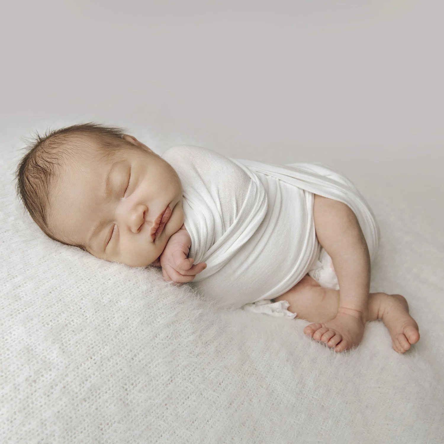 A sleeping newborn baby wrapped in a white blanket, lying on a soft white surface.