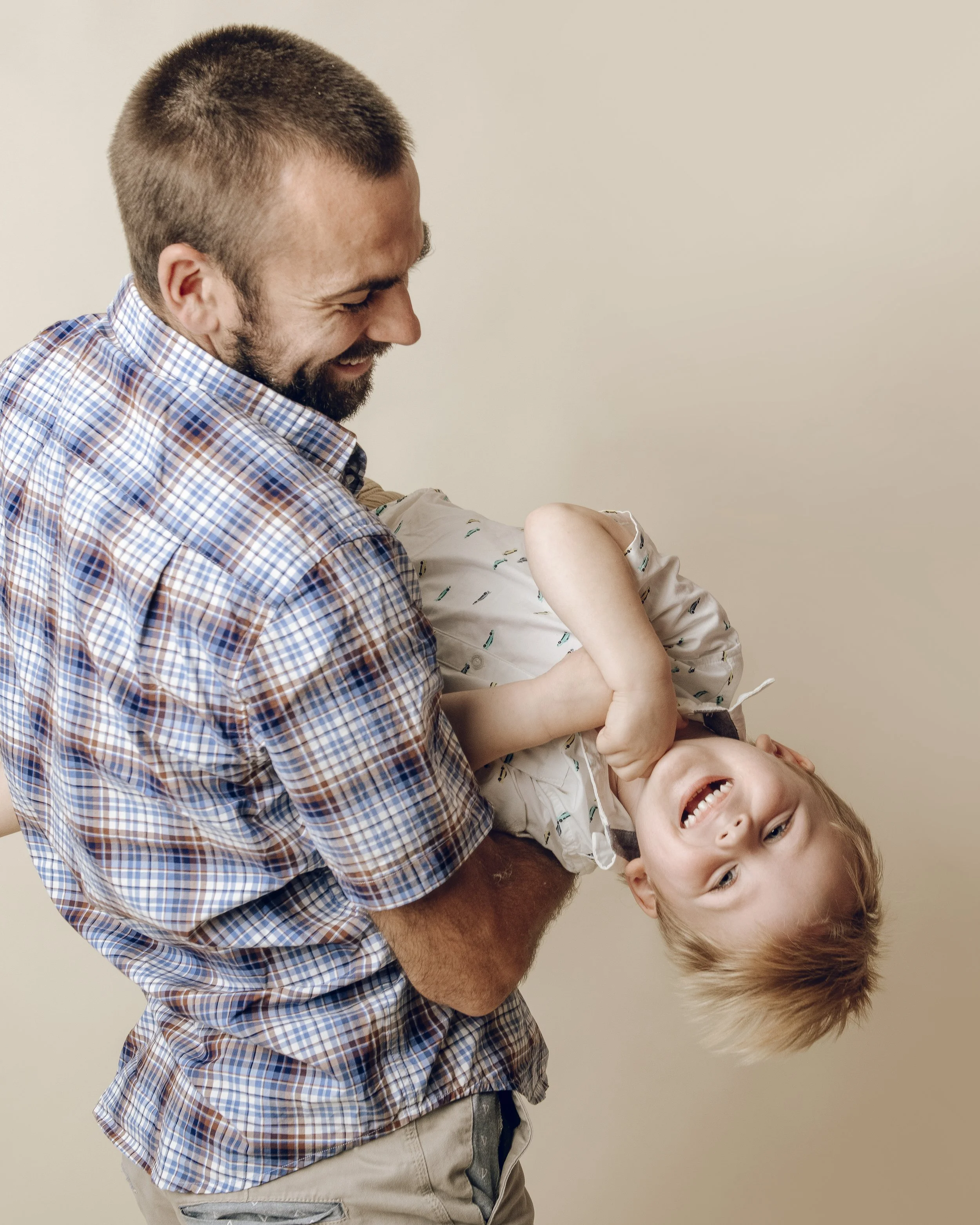 A man with a beard and short hair, wearing a checkered shirt, laughing while holding a smiling young boy in his arms, who is also smiling and wearing a light-colored shirt with small patterns, against a plain beige wall background.