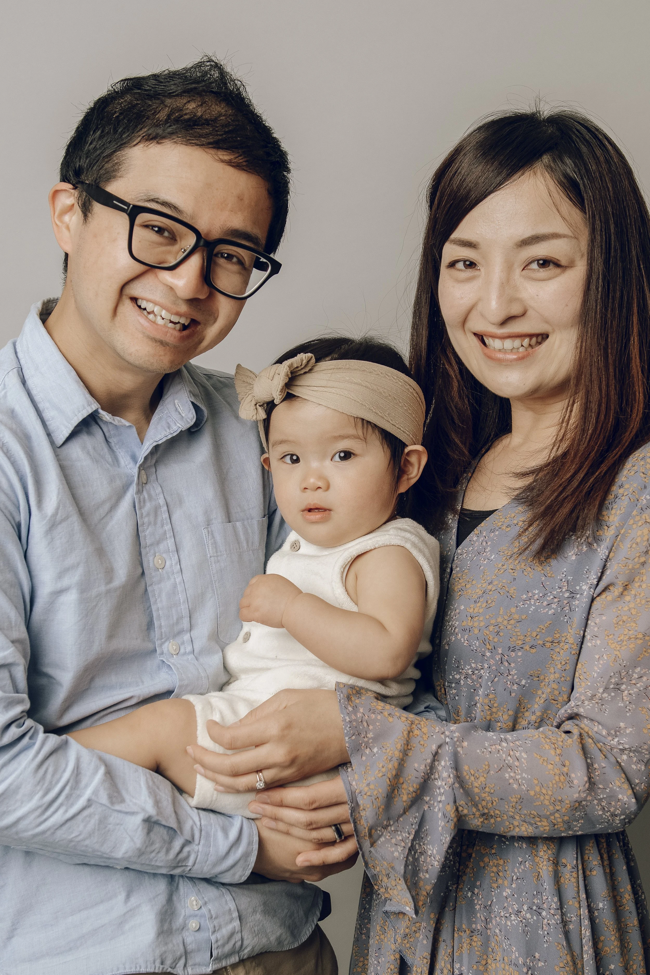 A family of three, including a man, woman, and young girl, smiling and posing together against a plain background. The man has glasses and is wearing a light blue shirt, the woman has long brown hair and is wearing a patterned dress, and the girl is wearing a beige headband and a white outfit.
