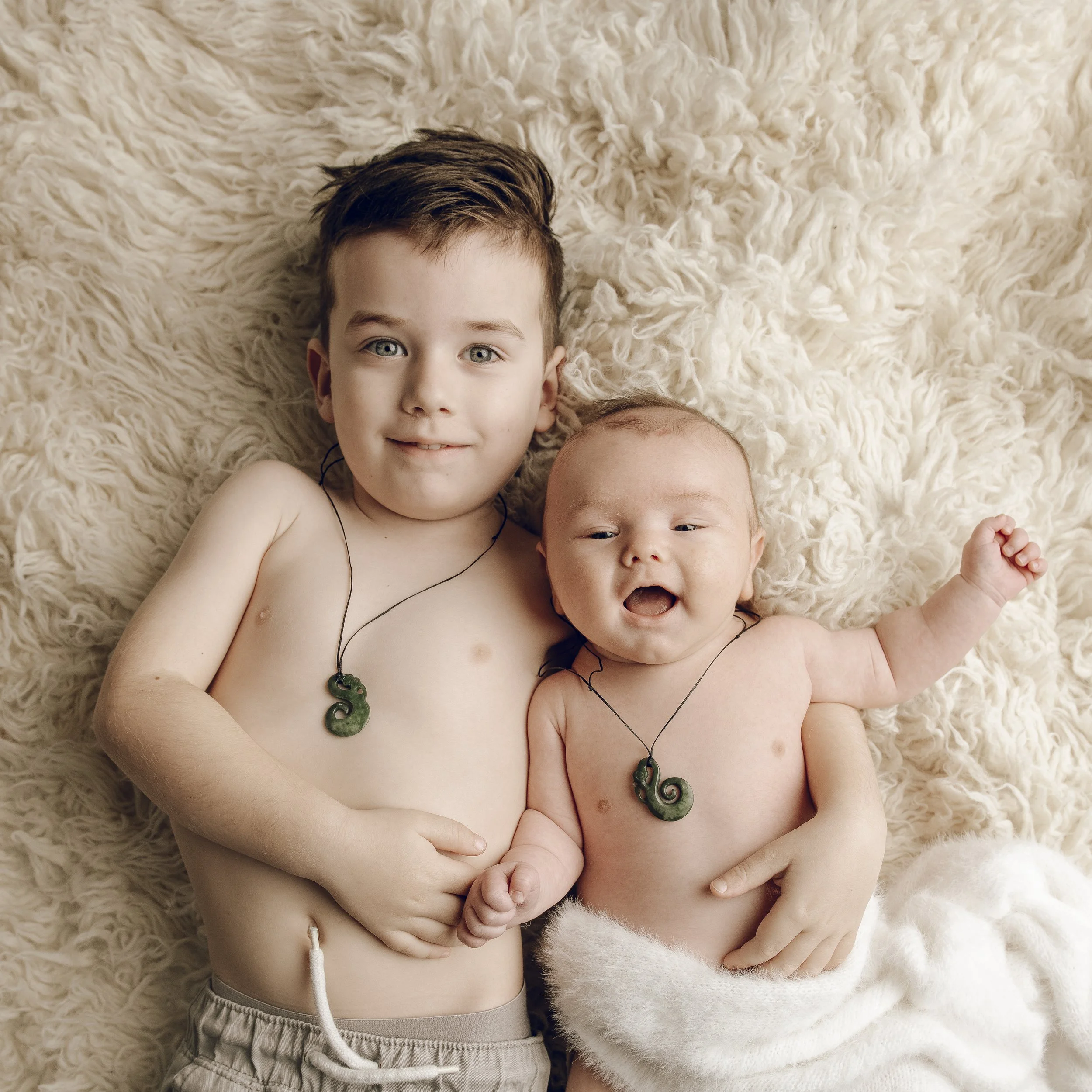 Two young children, a boy with blue eyes and brown hair and a girl, lying on a fluffy cream-colored blanket, both wearing necklaces with jade or green stone pendants.