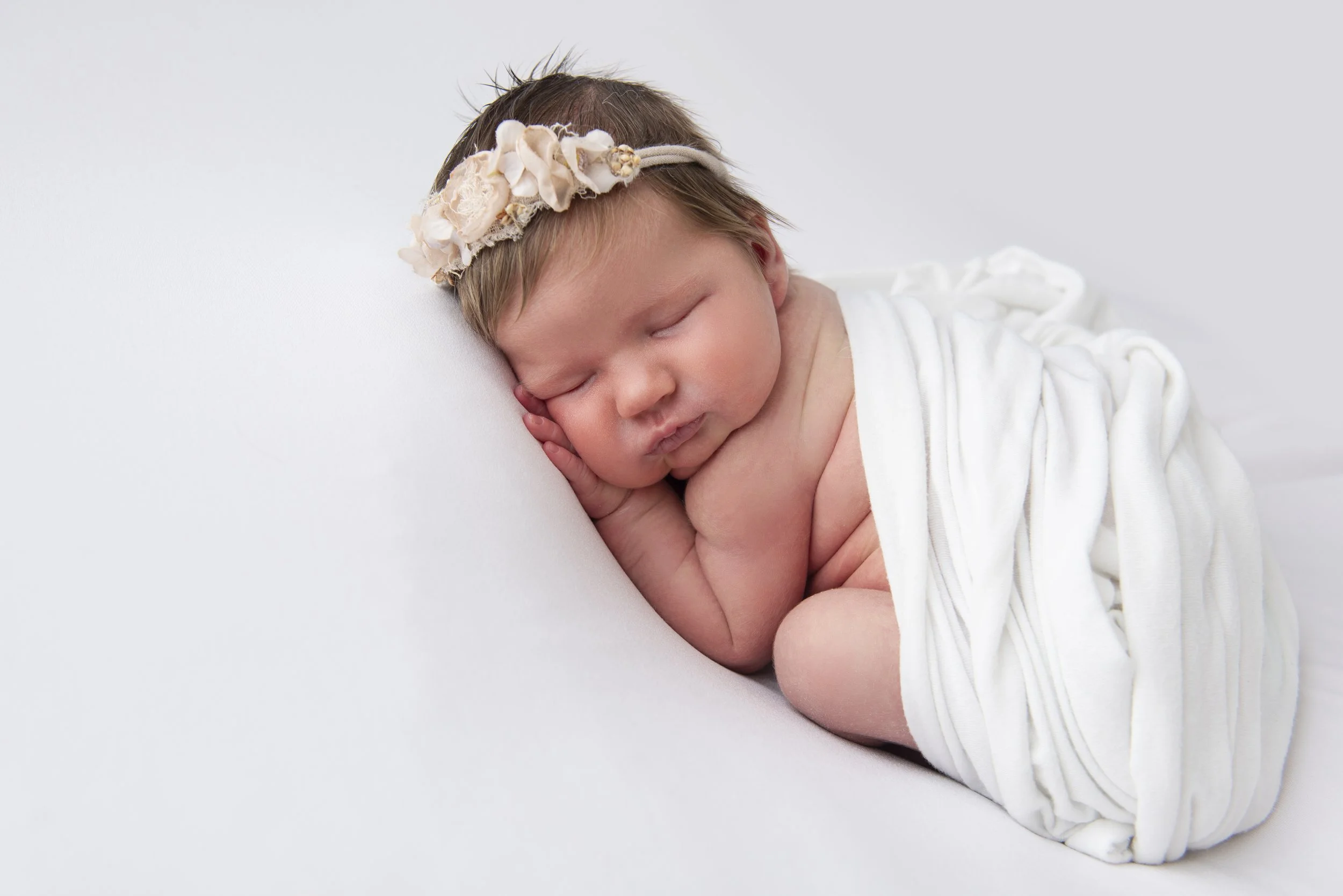 A sleeping baby girl lying on her side on a white surface, wearing a floral headband and a white outfit.