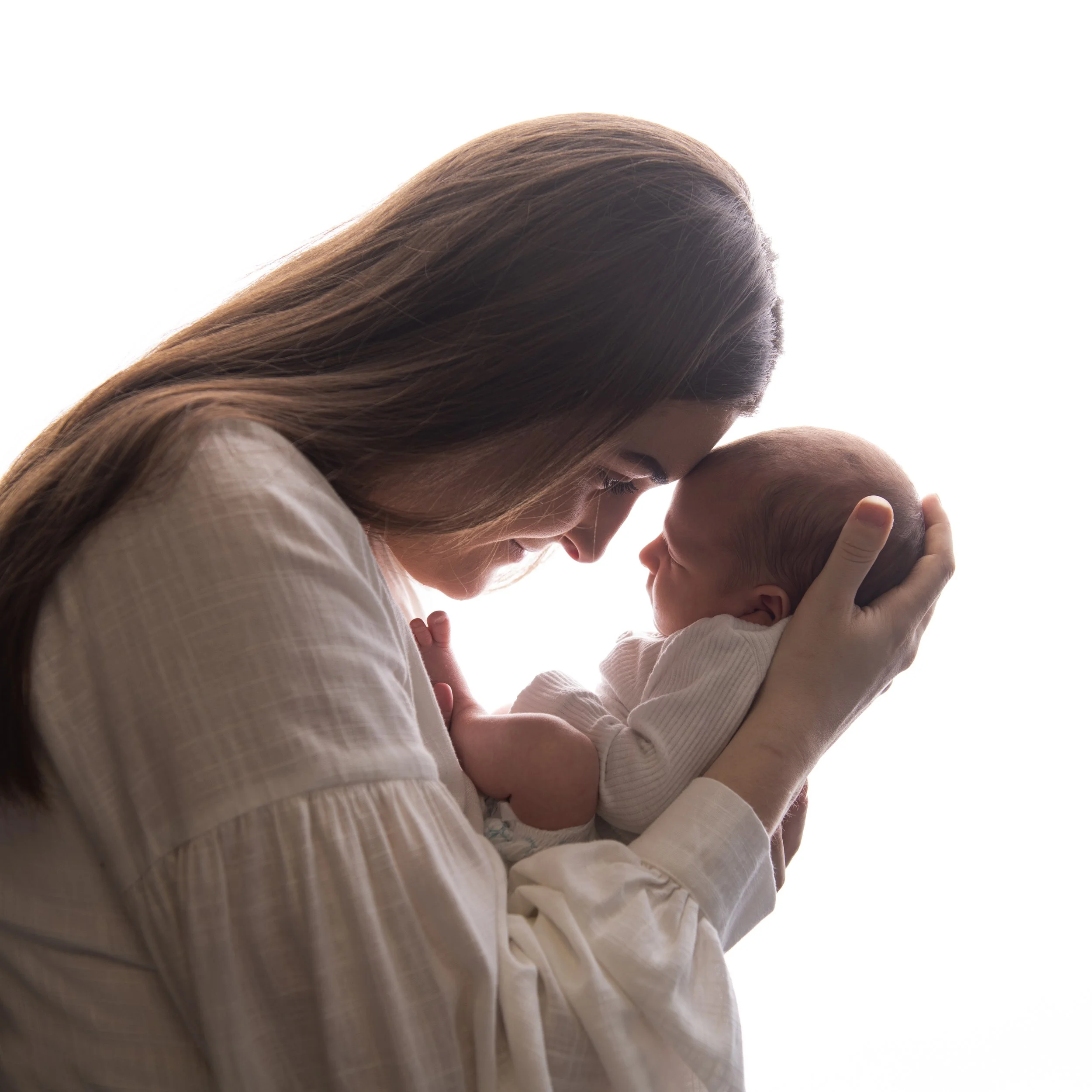A woman with long brown hair gently holding a baby in her arms, both looking at each other, against a white background.