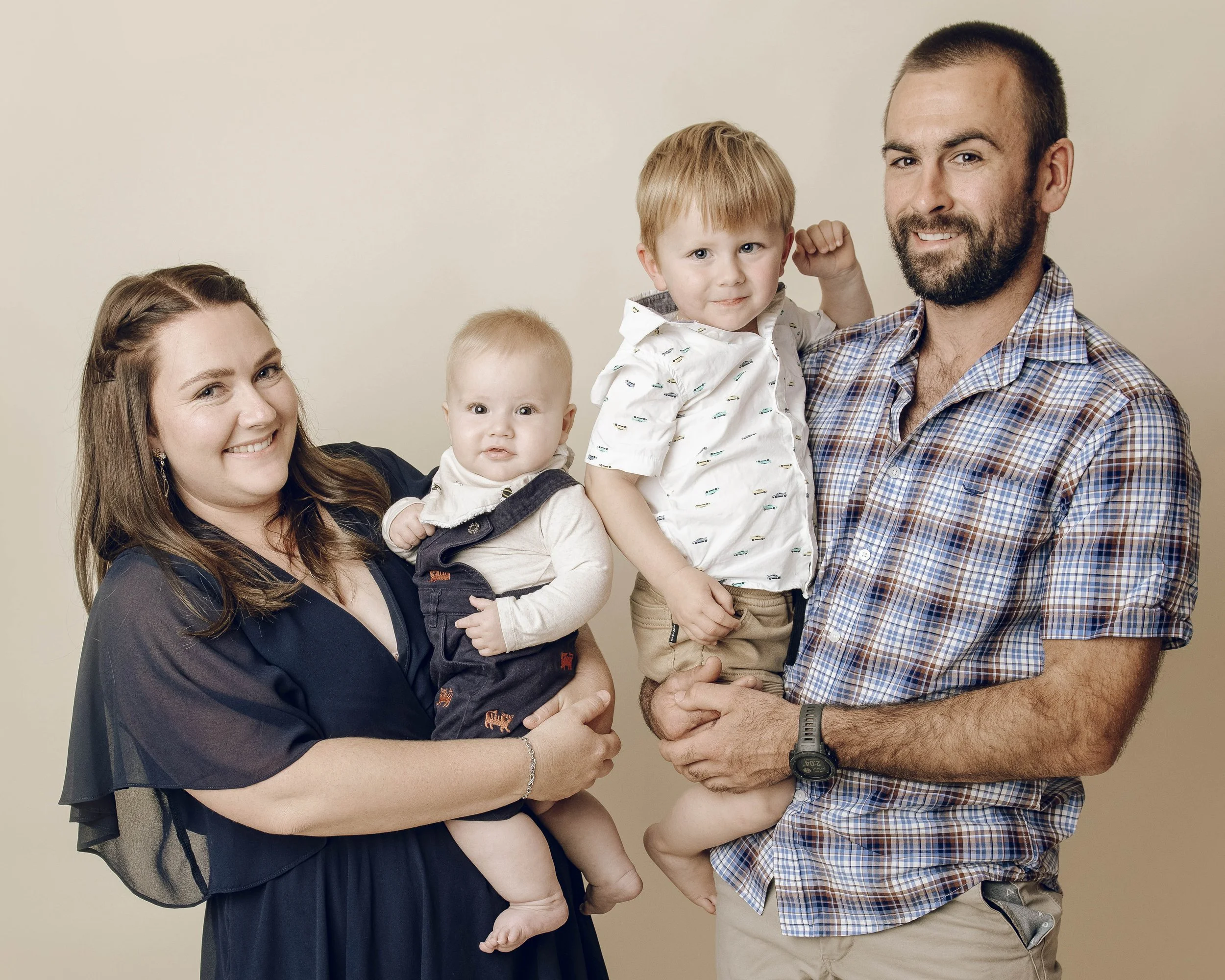 A smiling family of four, including a woman, a man, and two young children, posing against a plain beige background.