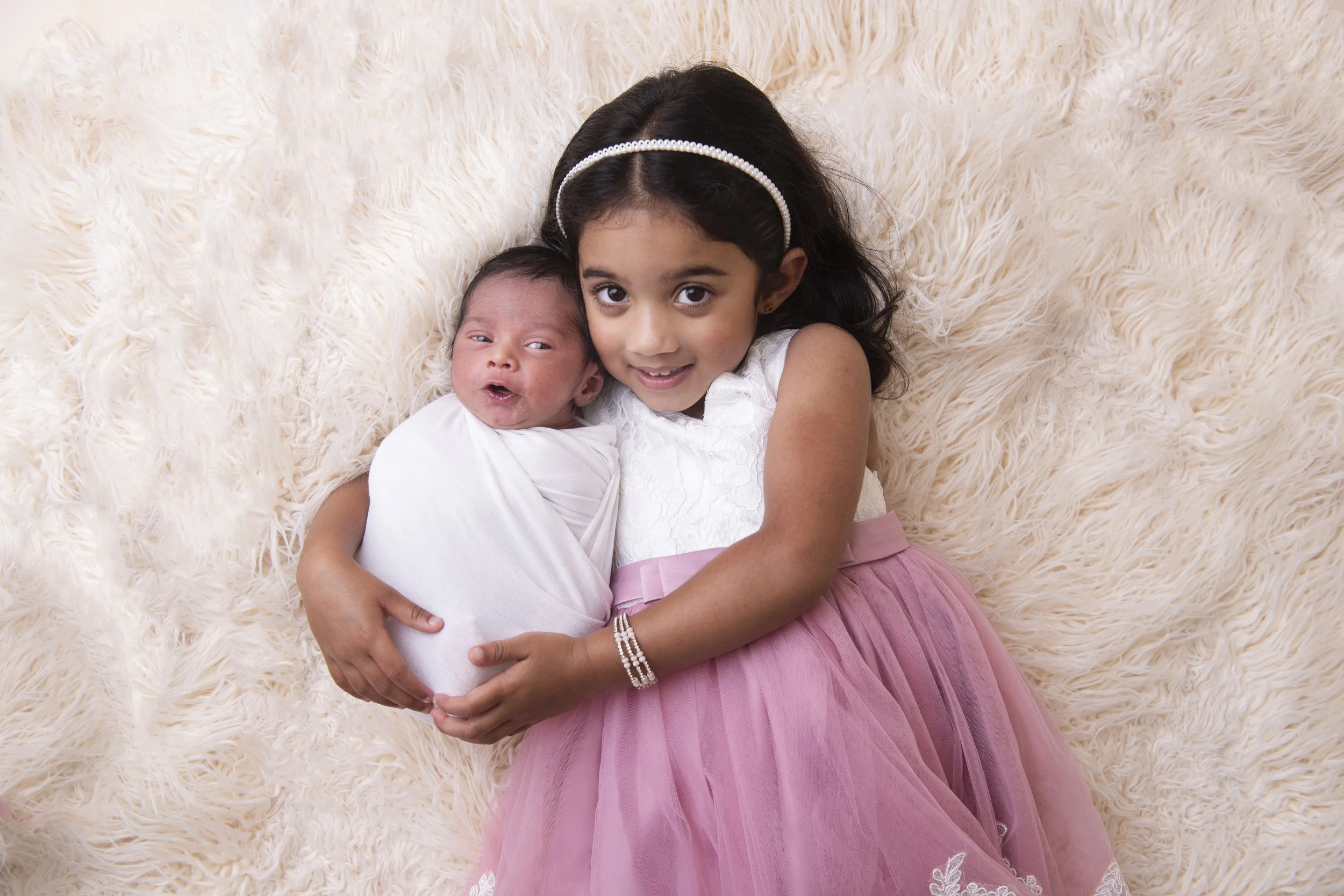 A young girl with dark hair, wearing a pearl headband and a white dress, holding a newborn baby with dark hair, dressed in a white outfit, lying on a fluffy cream-colored blanket.