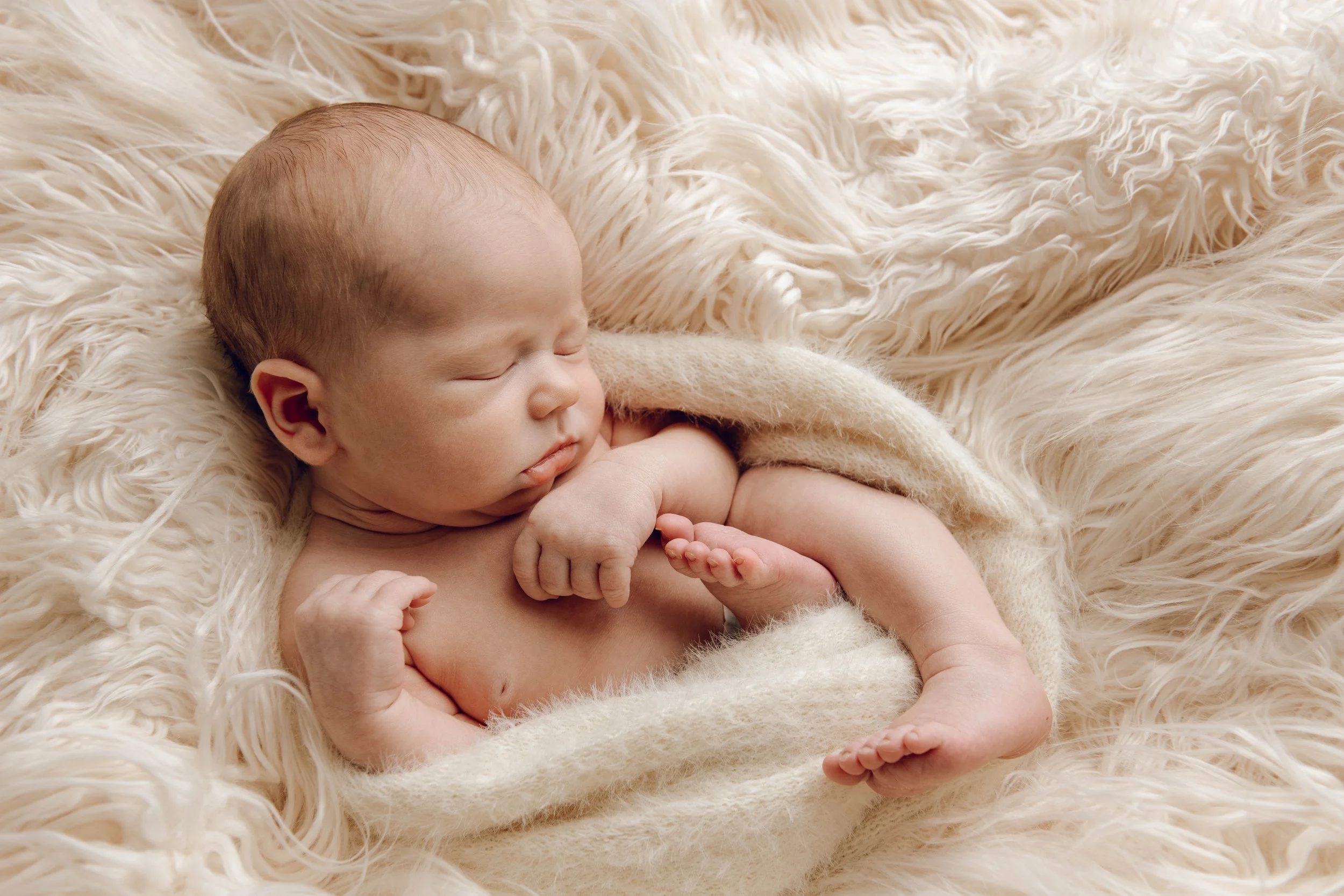 A sleeping newborn baby wrapped in a soft beige blanket, lying on a fluffy white rug.