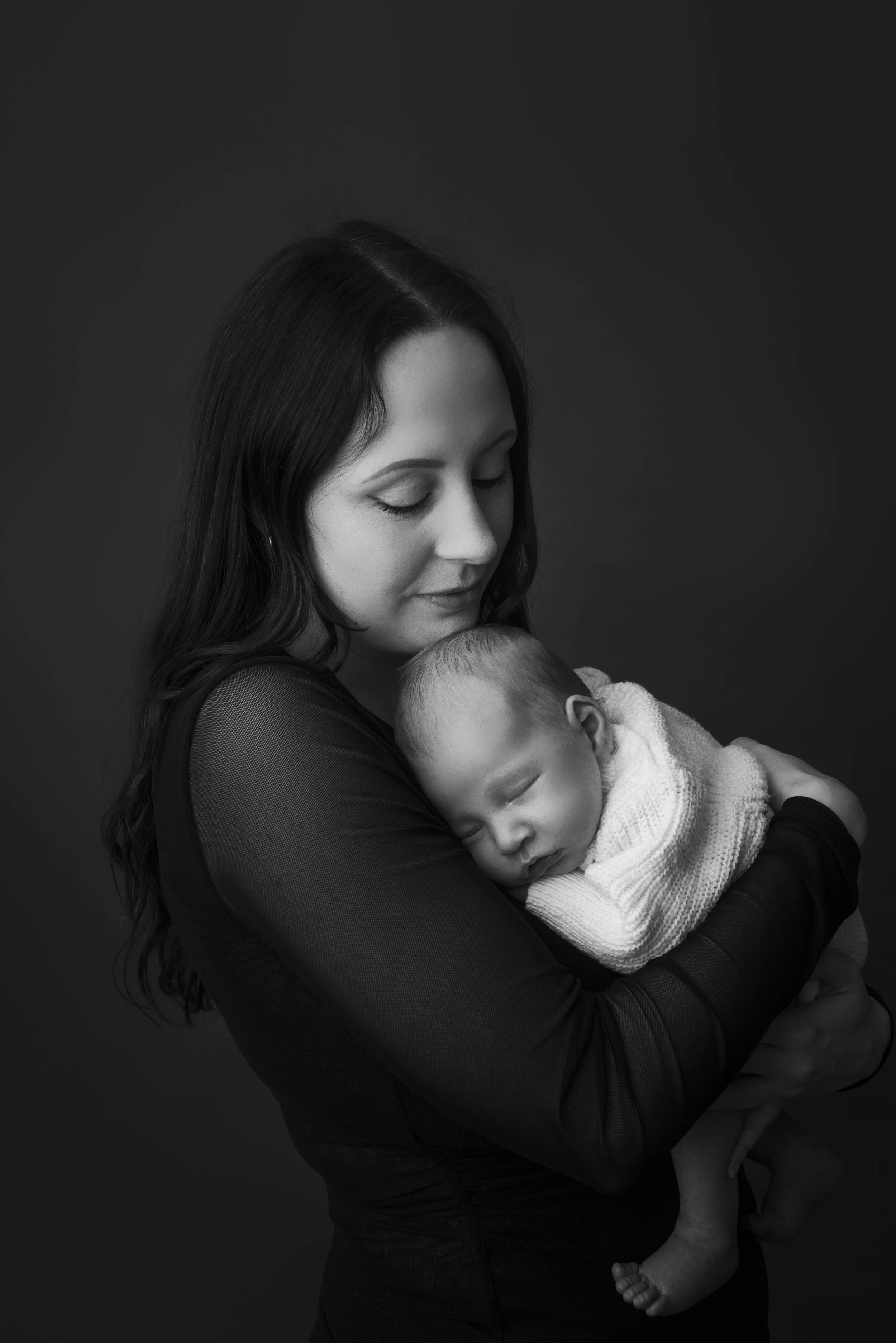 A woman with dark hair and closed eyes holds a sleeping baby in her arms against a dark background.