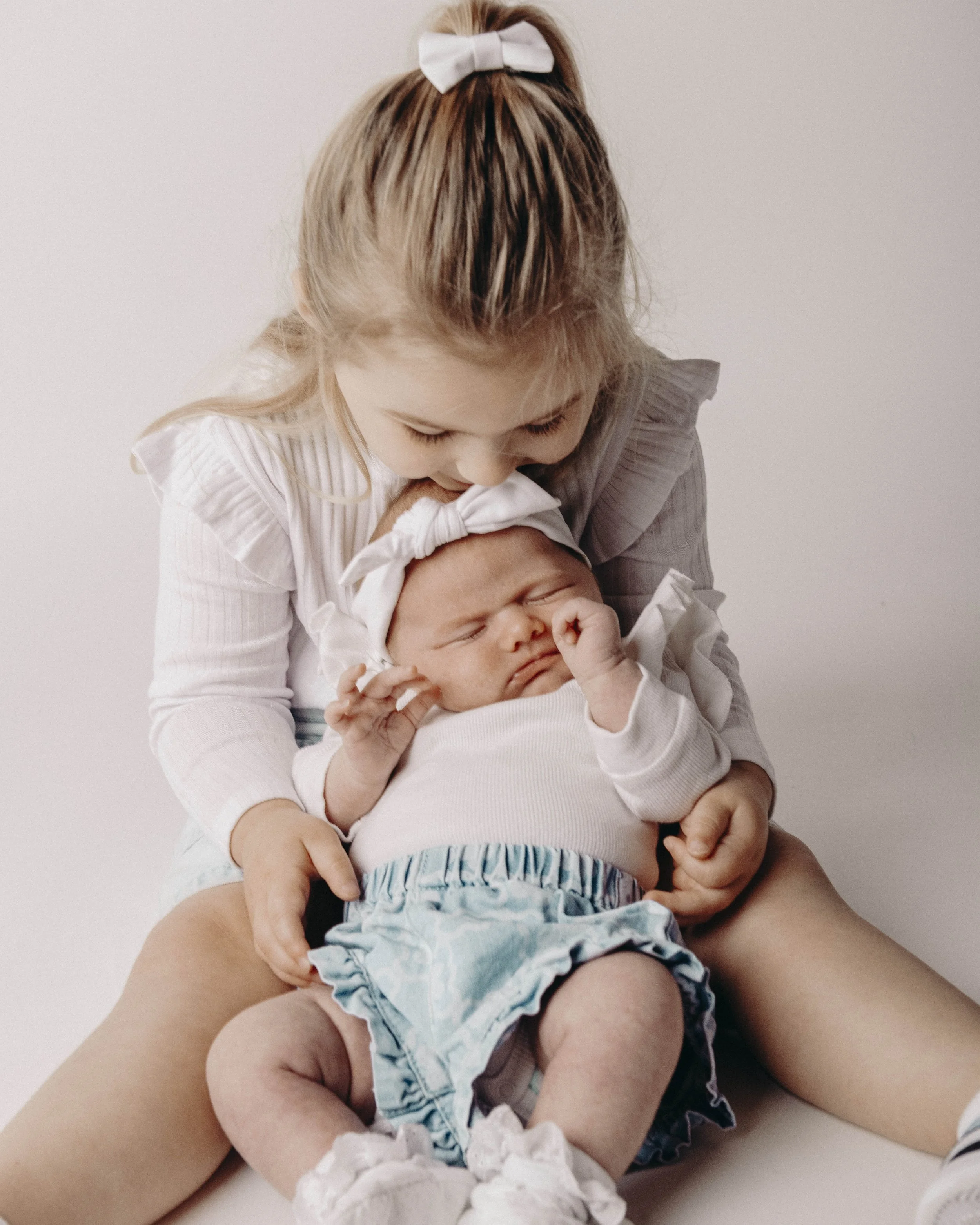 A woman holding a sleeping baby in her arms while sitting on the floor, with the woman’s face bowed and eyes closed, wearing a white shirt with ruffled sleeves and a white bow in her hair, against a plain white background.