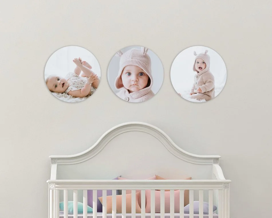 Three circular photo frames of babies on a wall above a white crib in a nursery, with colorful pillows inside the crib.