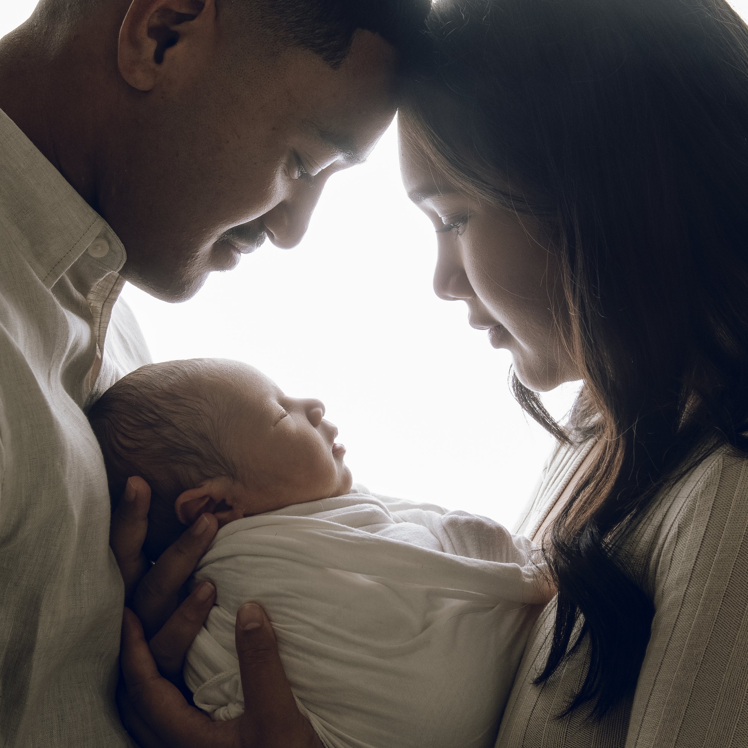 A close-up of a family with two adults and a baby, showing their faces leaning towards each other with their eyes closed, in an intimate and tender moment.