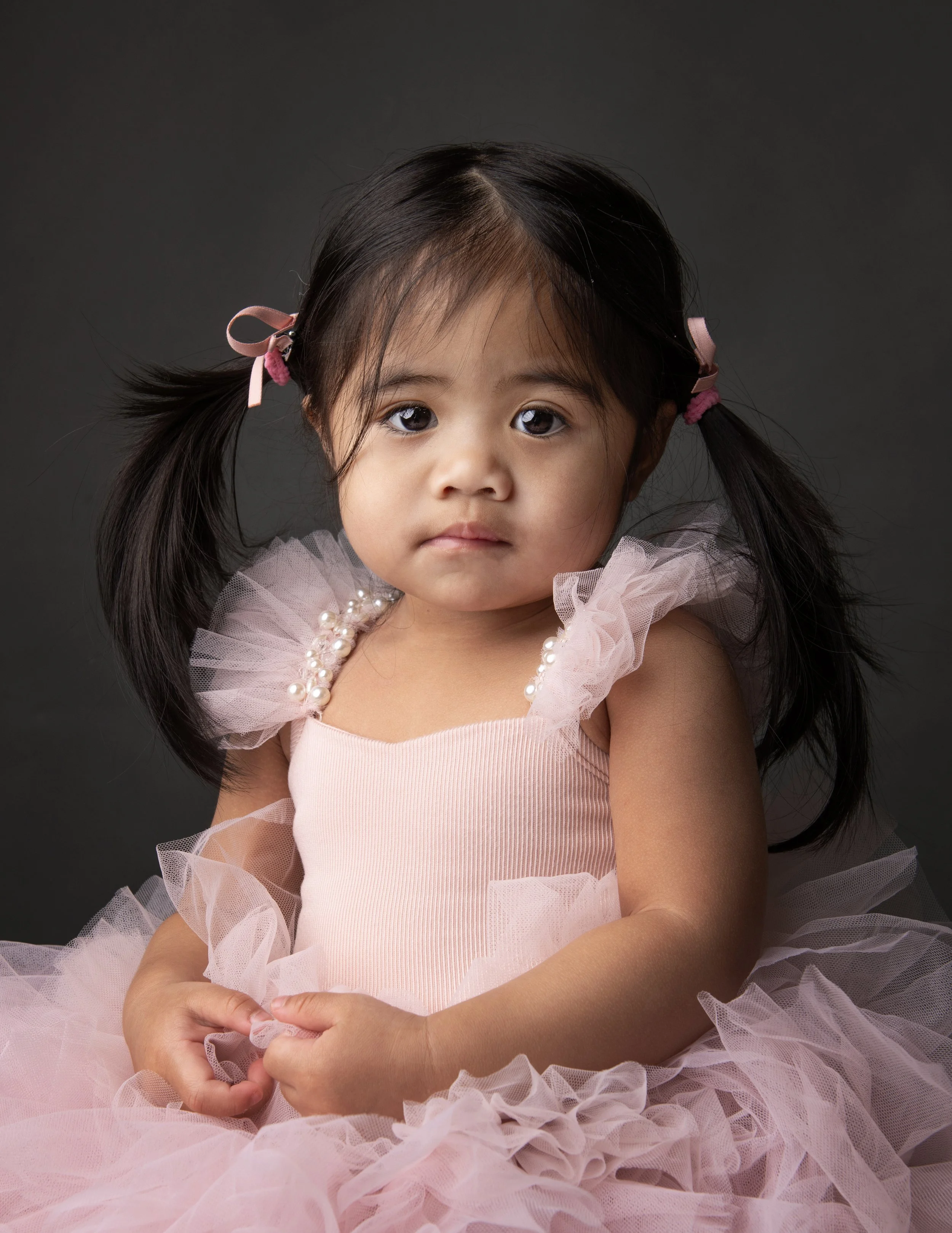 A young girl with dark hair styled in pigtails, wearing pink ribbons and a pink dress with ruffled tulle sleeves and pearl accents, against a dark background.