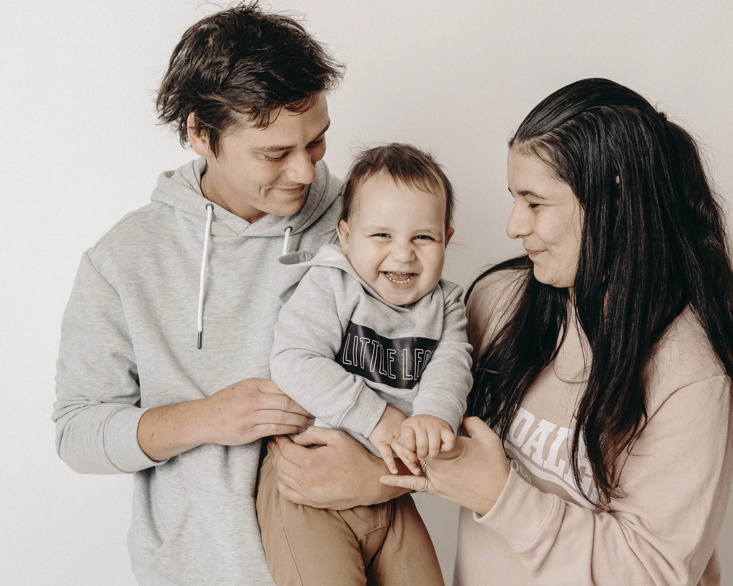 A family of three, including a man, woman, and toddler, sharing a happy moment together, smiling and hugging against a plain white background.
