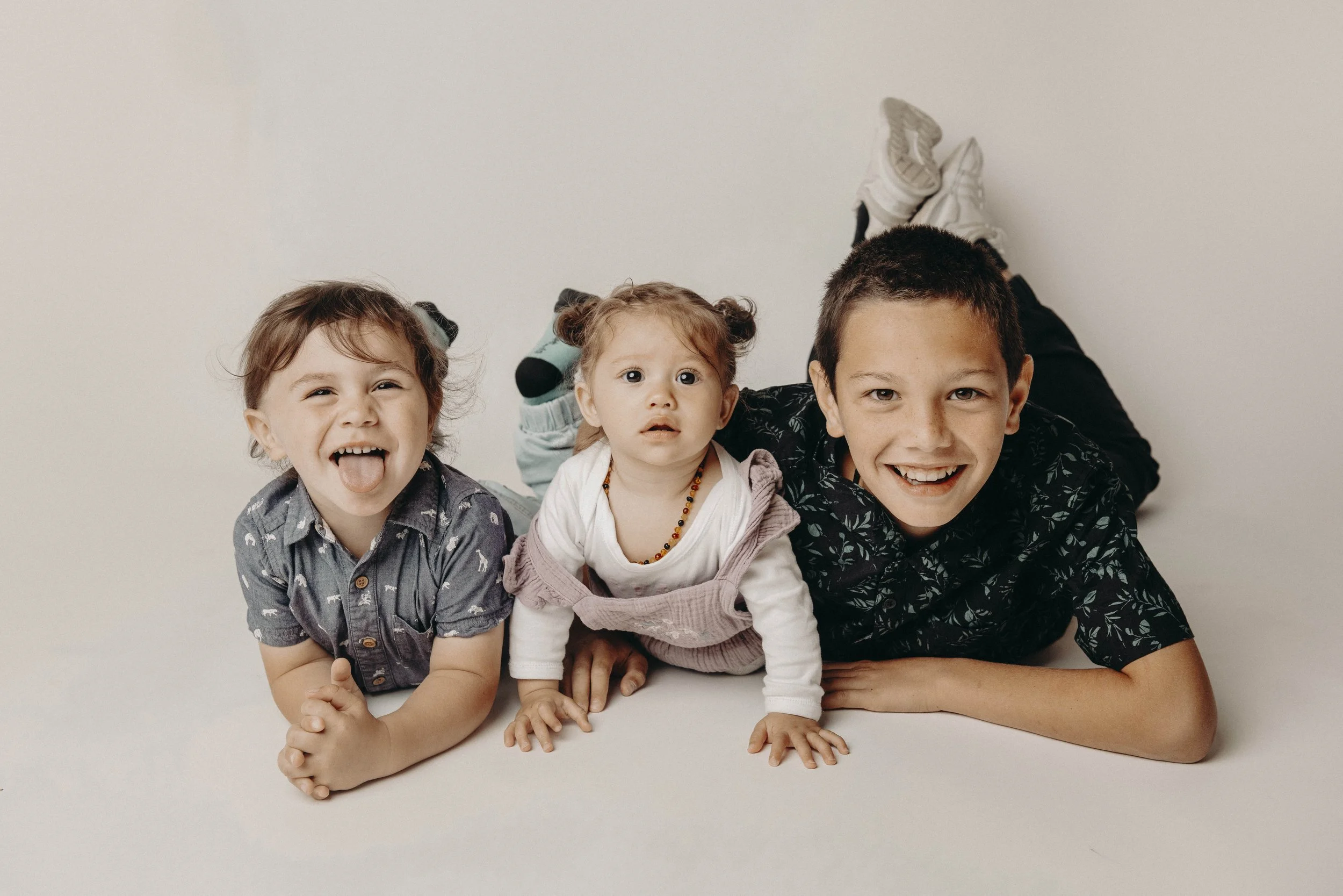 Three children lying on the floor with their heads resting on their arms, smiling and having fun against a plain white background.