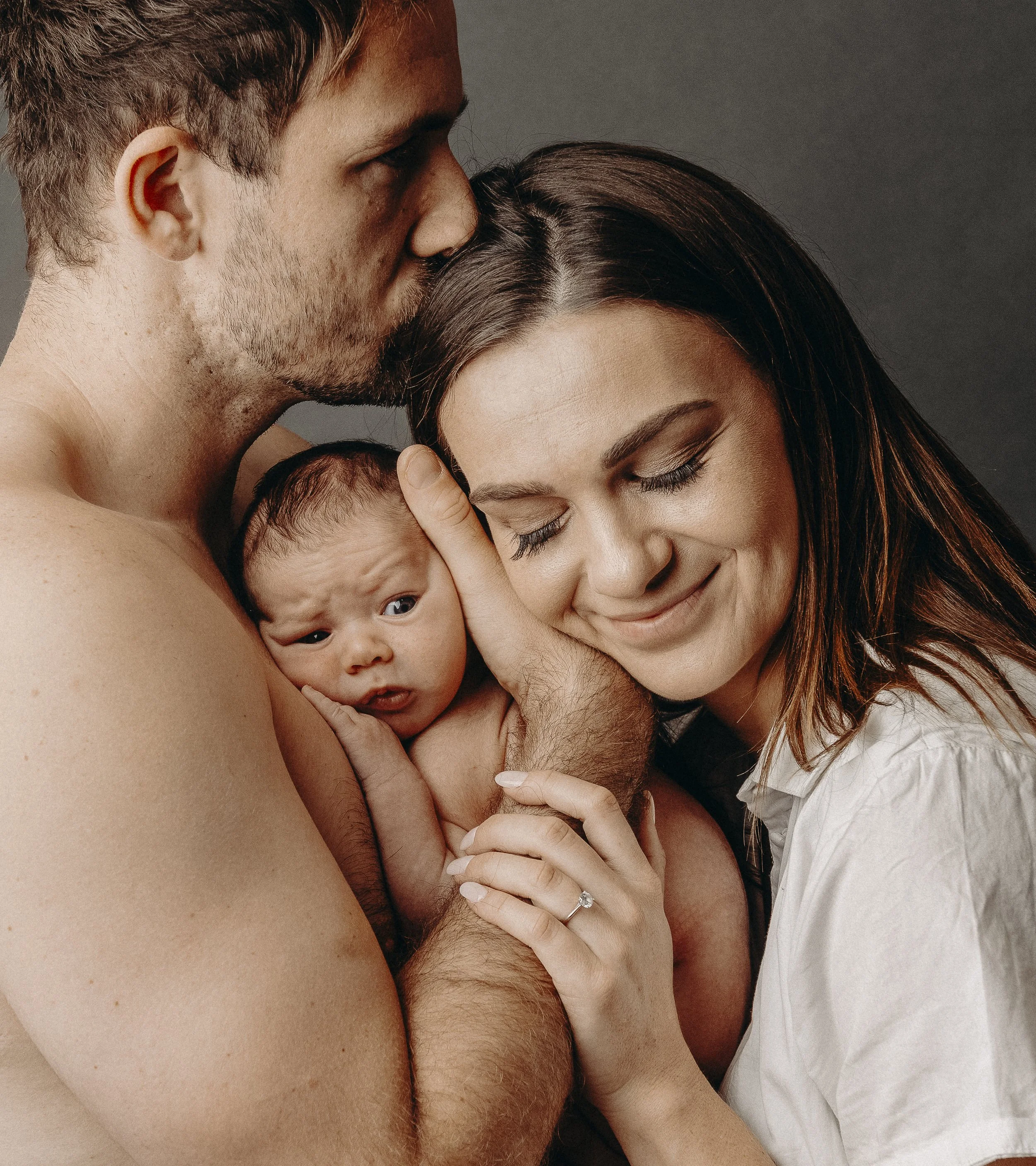 A family of four, including a man, woman, and two children, sharing an intimate moment of closeness and affection. The man is kissing the woman on the forehead, and the woman is holding one of the children with a gentle smile.