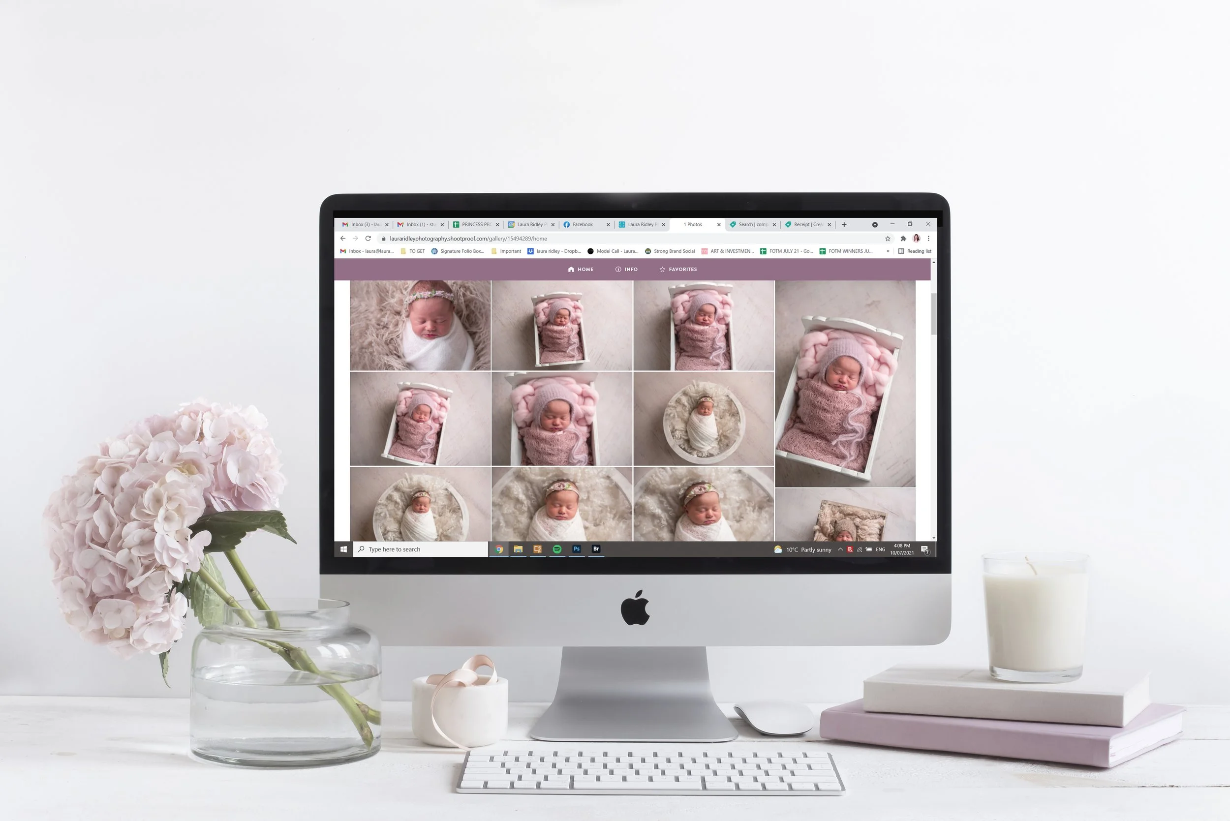 Computer monitor displaying photographs of a newborn baby, with a pink theme. The monitor is on a white desk with pink hydrangea flowers in a glass vase, a small white candle, a white keyboard, and a glass of milk nearby. The background is plain white.
