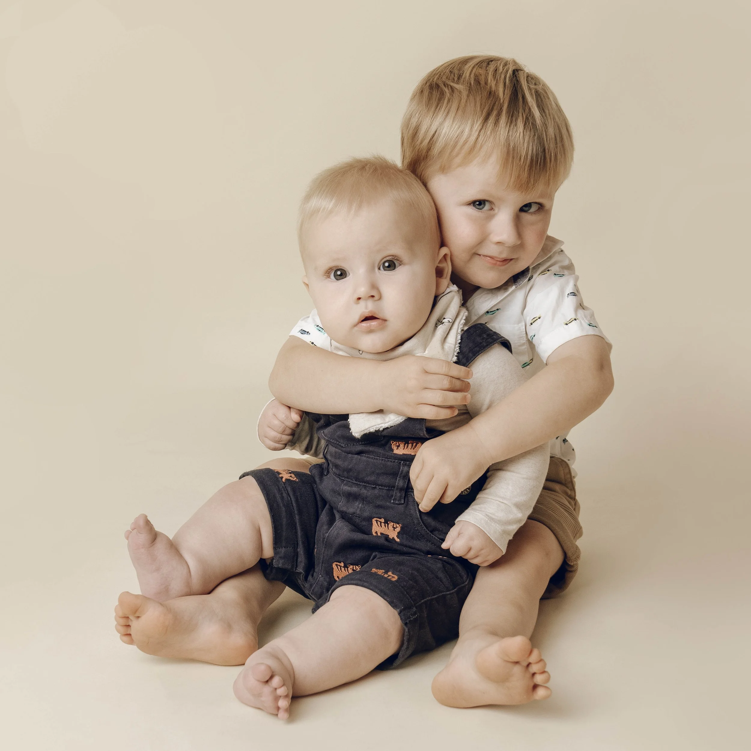 A young boy with blonde hair and blue eyes hugging a baby with blonde hair and blue eyes, both sitting on a beige background.