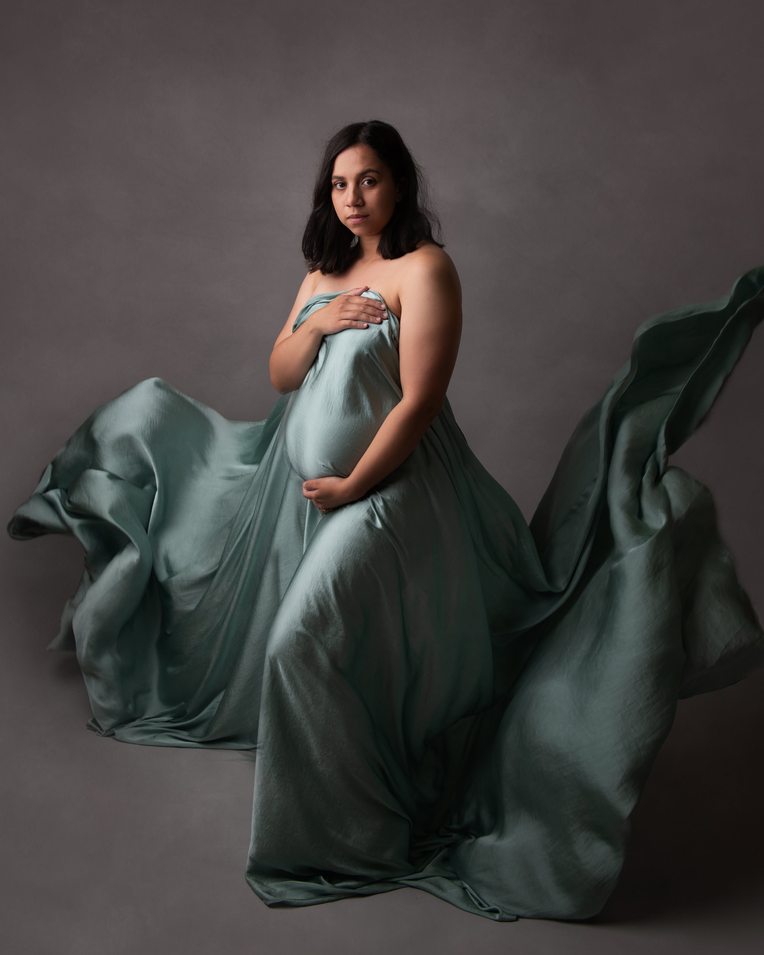 A woman with medium-length dark hair and a serious expression, draped in a flowing, shiny, green satin fabric, stands against a neutral gray background.