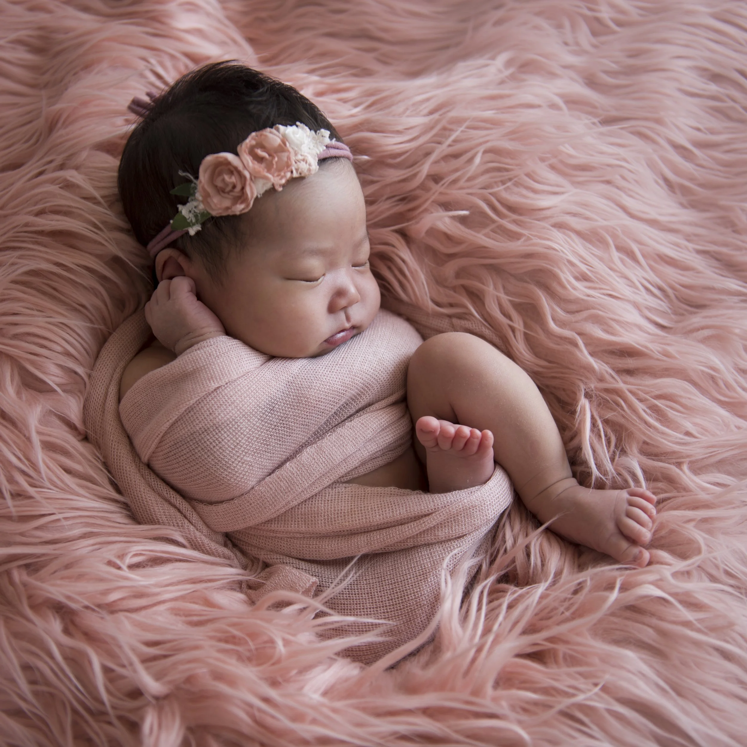 A sleeping baby with a floral headband, lying on a pink furry blanket.