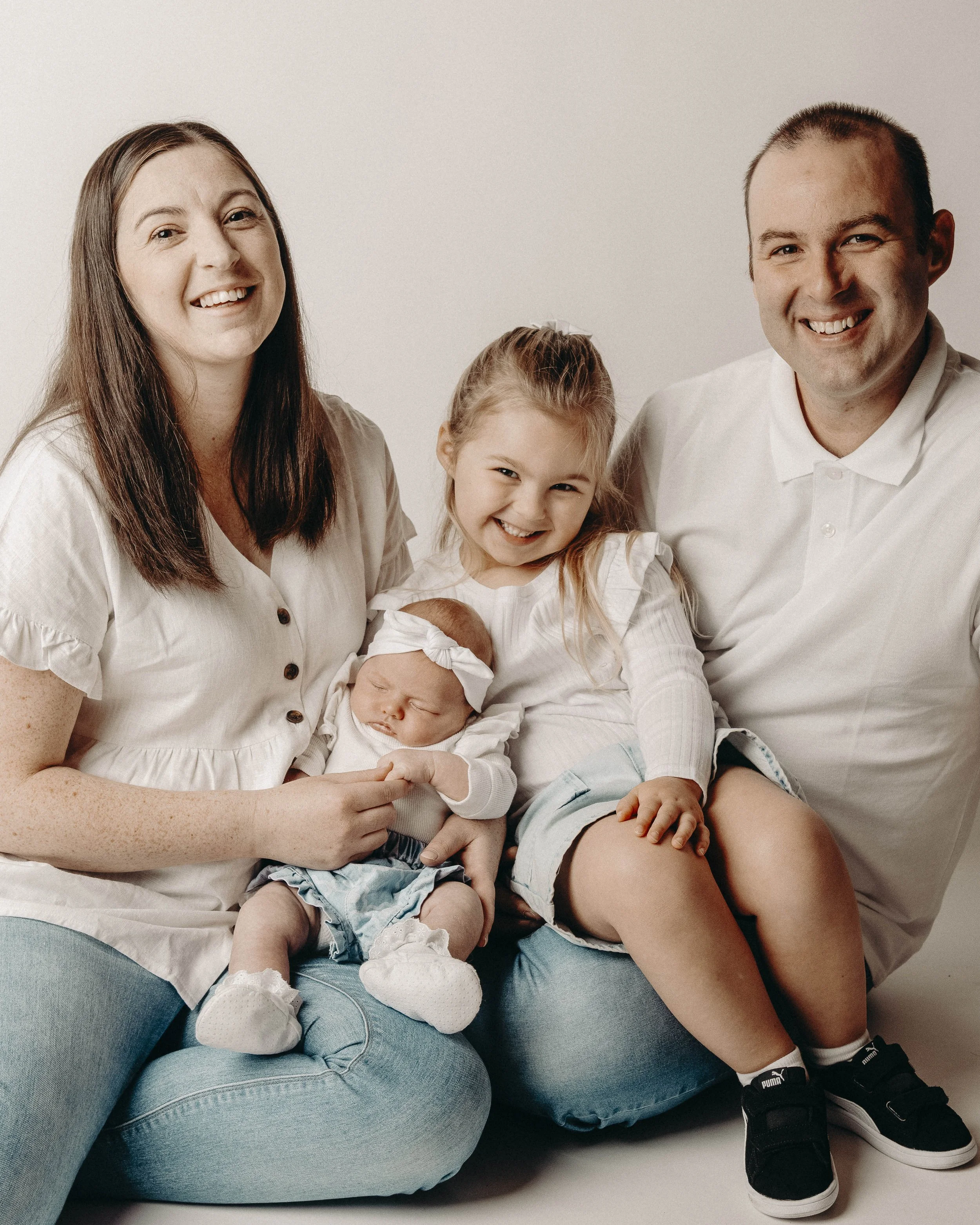 Family portrait of four with two women, a man, a girl, and a baby, sitting on a blue couch against a white background. All are smiling or content, with the girl making a playful face and the baby sleeping.