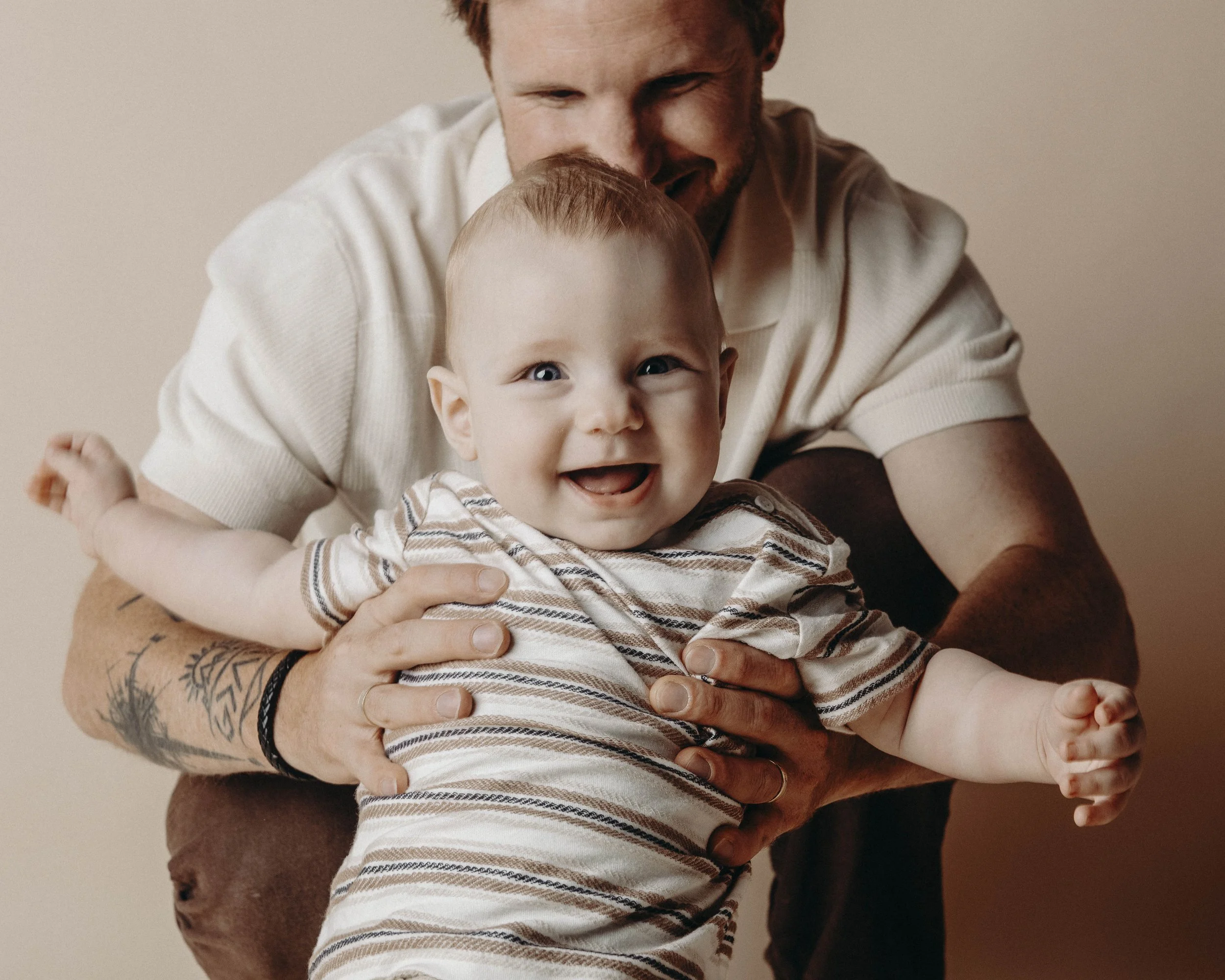 A man with tattoos and a beige shirt holding a smiling baby with blue eyes and a striped shirt, against a plain beige background.