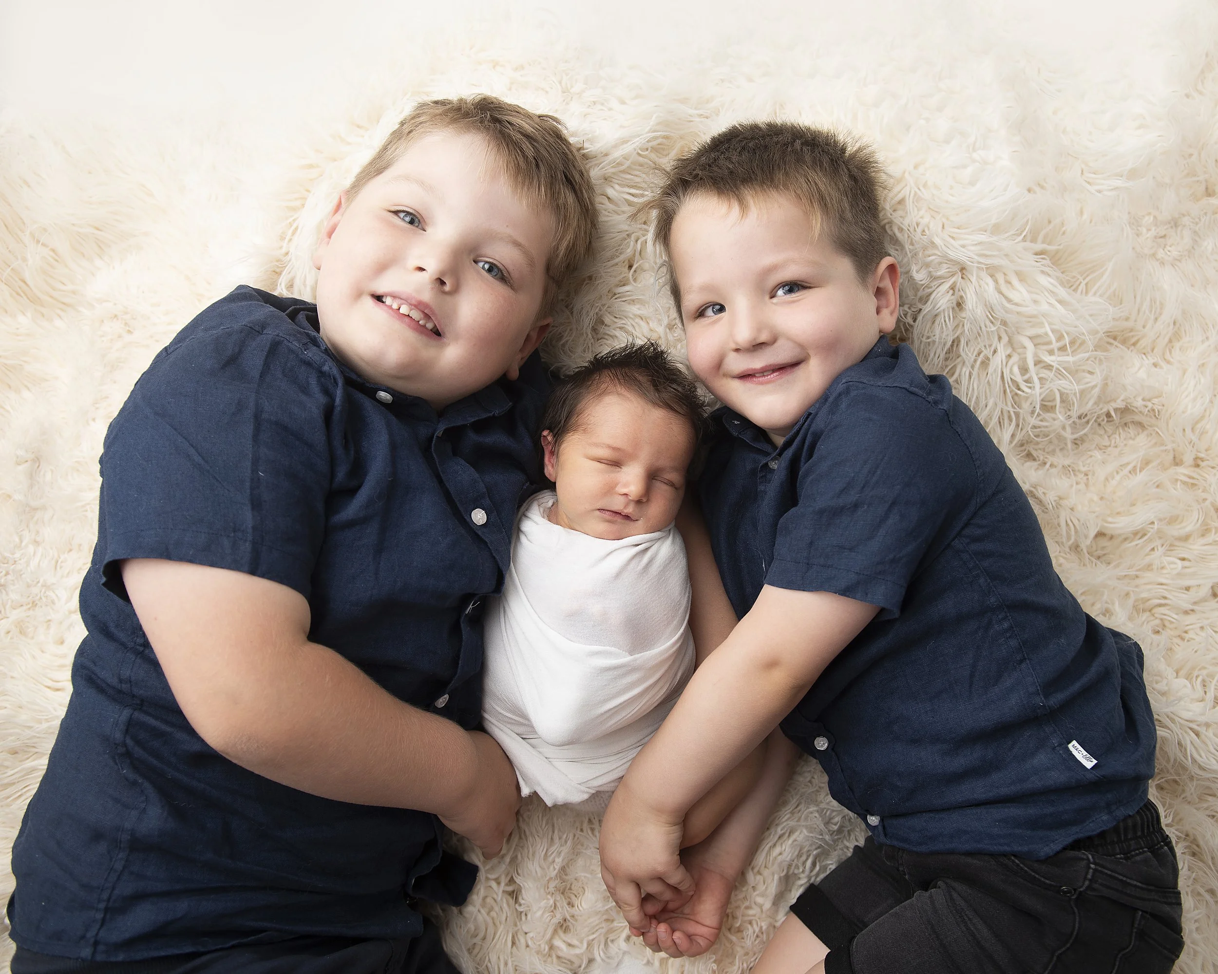 Four children lying on a fluffy cream-colored rug, smiling and hugging each other.