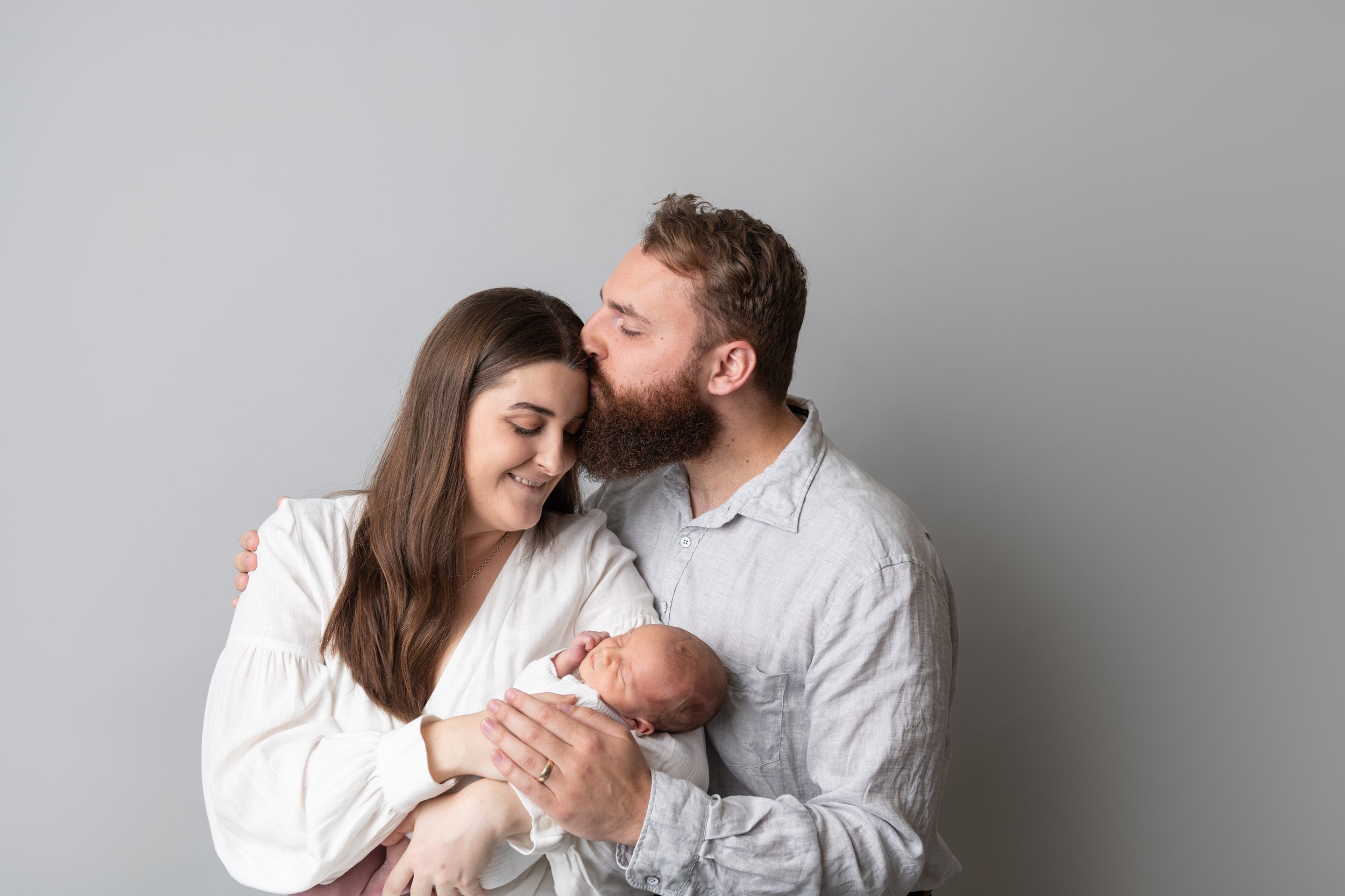 A couple holding a newborn baby, with the man kissing the woman's forehead while she smiles, against a plain gray background.